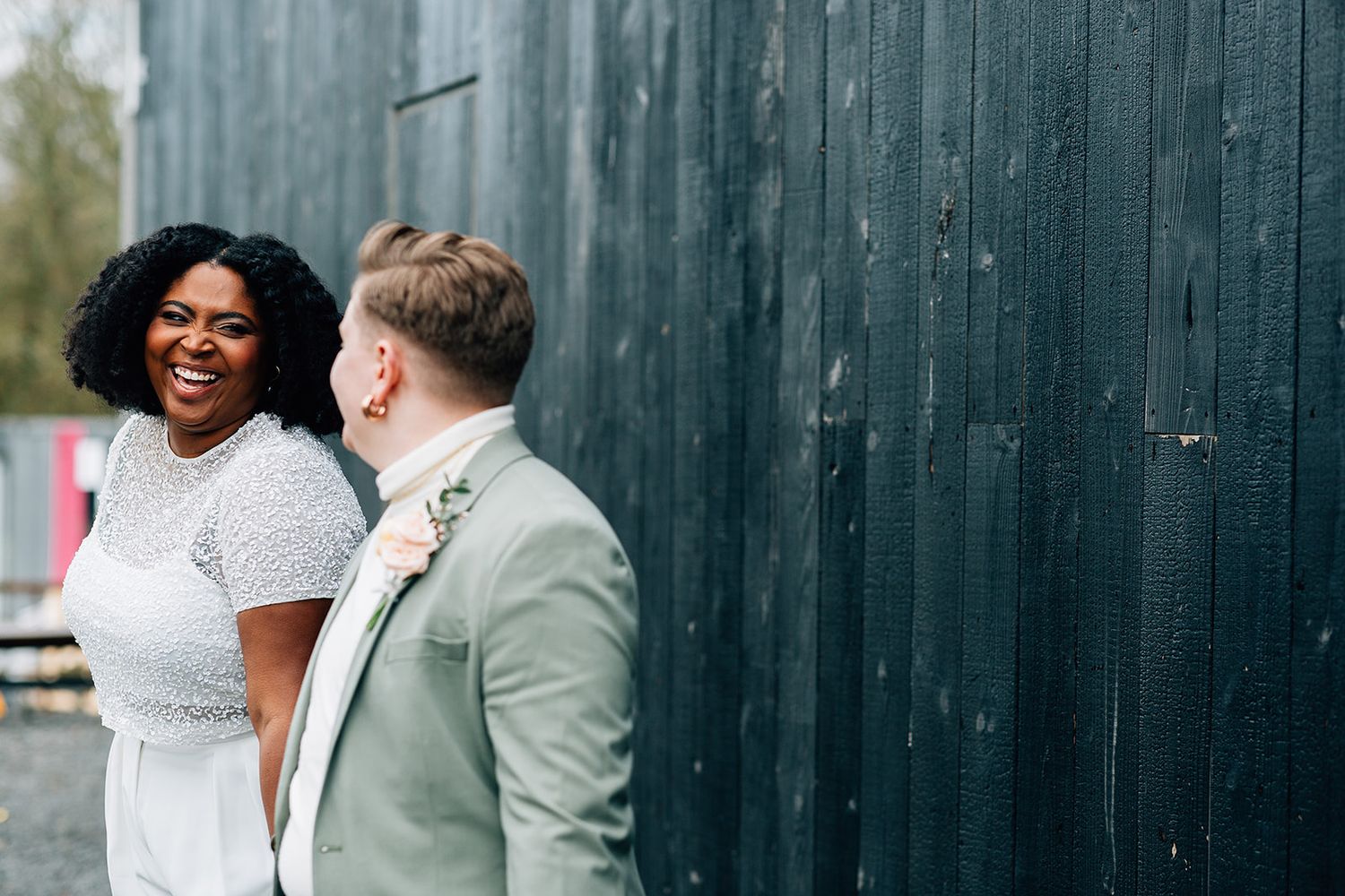 A couple shares a joyful moment together outdoors against a dark wooden fence backdrop.