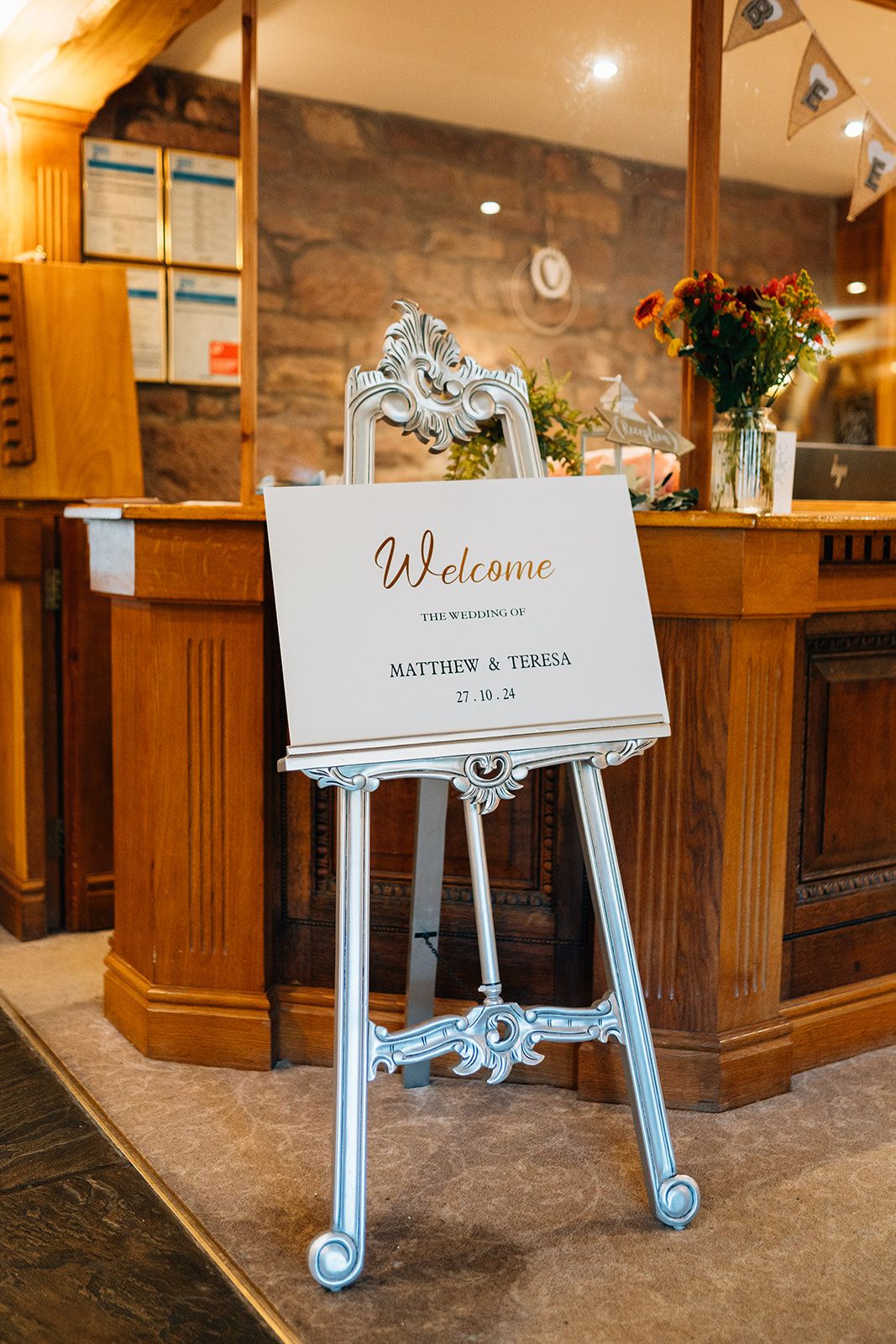 Wedding welcome sign for Matthew and Teresa on an ornate silver easel beside a wooden reception desk with a bouquet of autumn flowers.