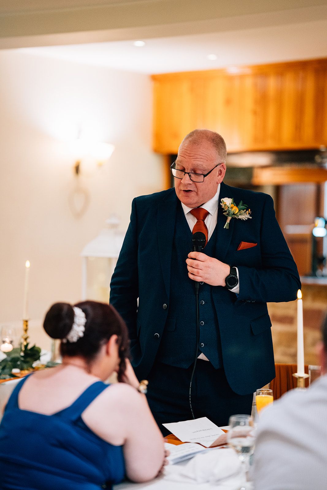 Man in a navy suit with a floral boutonniere gives a wedding speech into a microphone while a seated woman with a hair accessory listens.