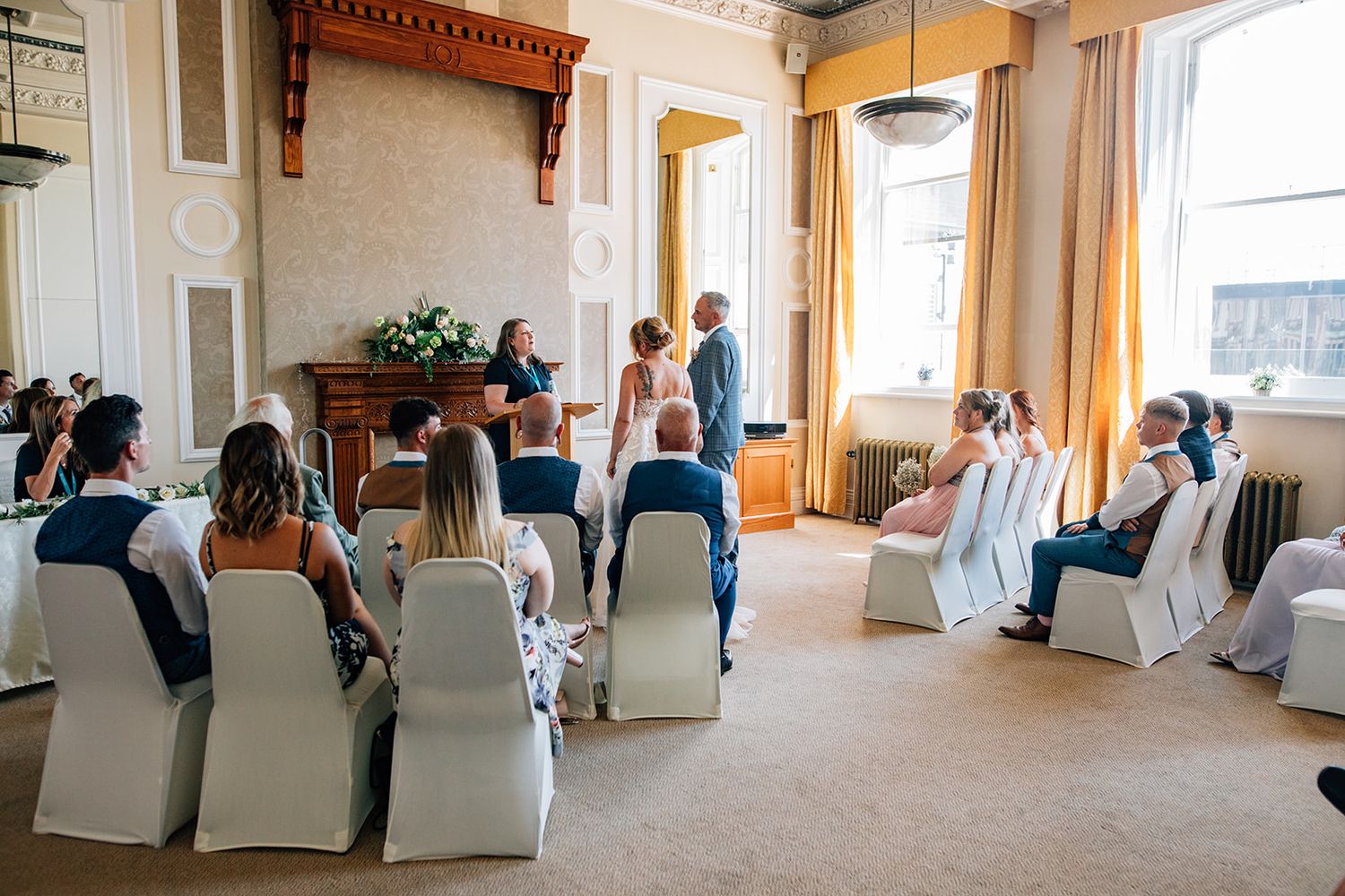 A panoramic view of a wedding ceremony venue with white chairs and large windows letting in natural light.