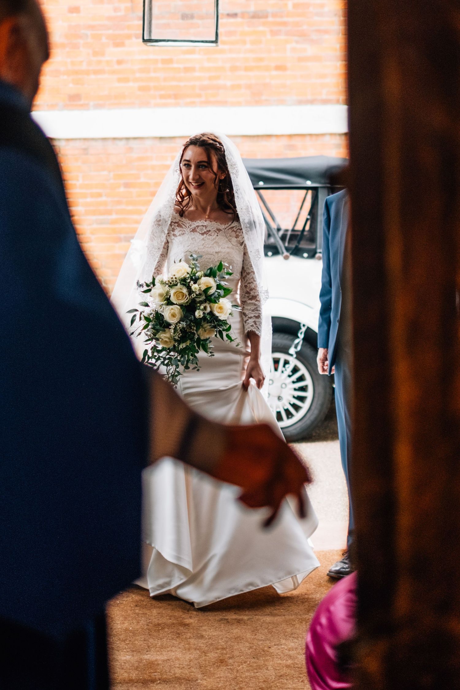 Smiling bride in a white lace gown and veil holding a white rose bouquet, framed by a doorway with a vintage car behind her.