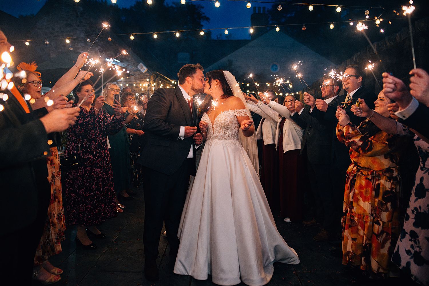 Wedding guests dance and celebrate under twinkling lights at an outdoor evening reception.