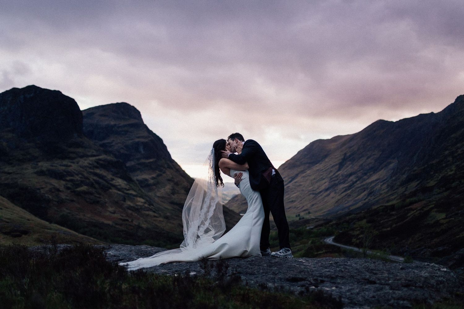 Bride and groom share a romantic kiss at dusk with dramatic Scottish Highland mountains in the background.