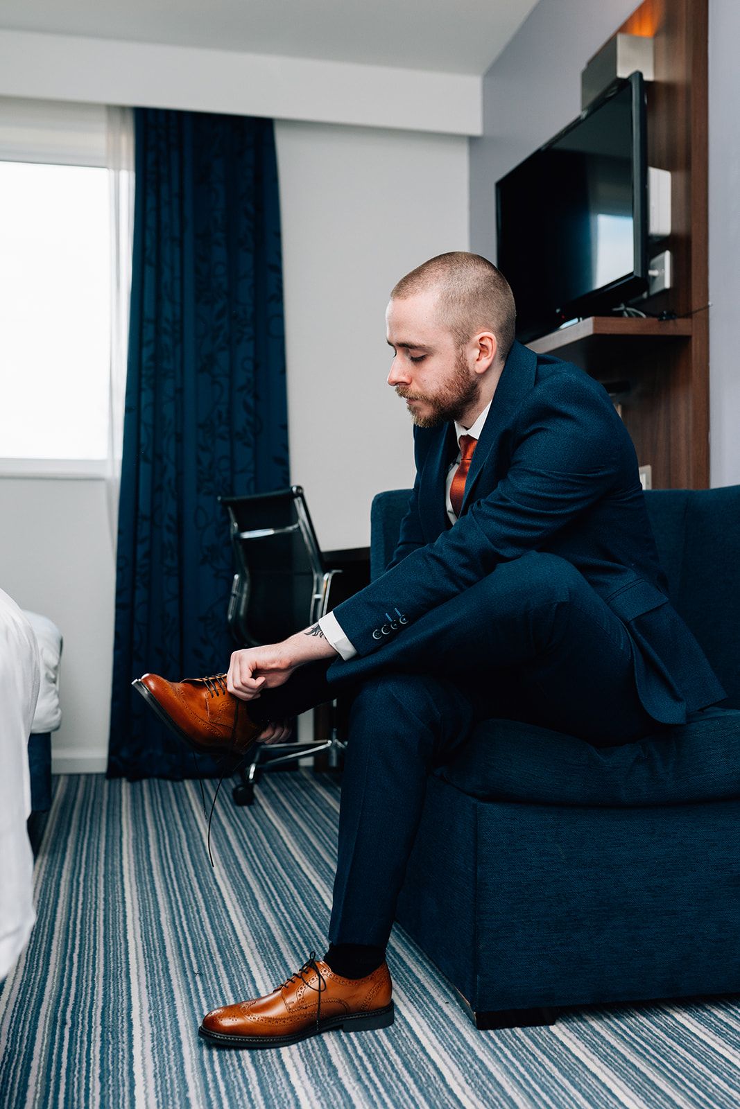 Man in a navy suit and red tie sits on a blue armchair in a hotel room, tying his brown leather dress shoe.