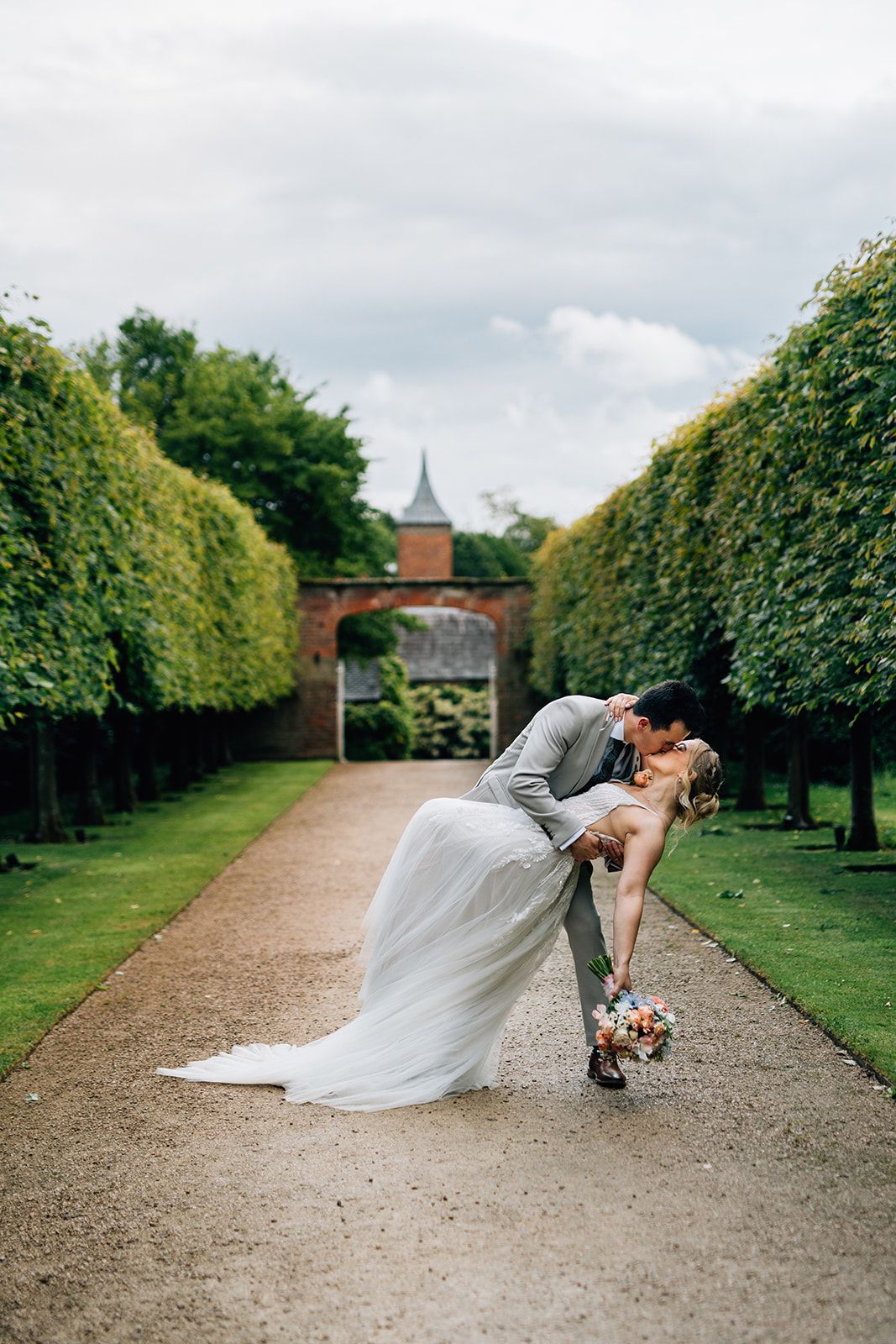 A romantic wedding moment captured in a formal garden path lined with manicured hedges and a church spire in the background.