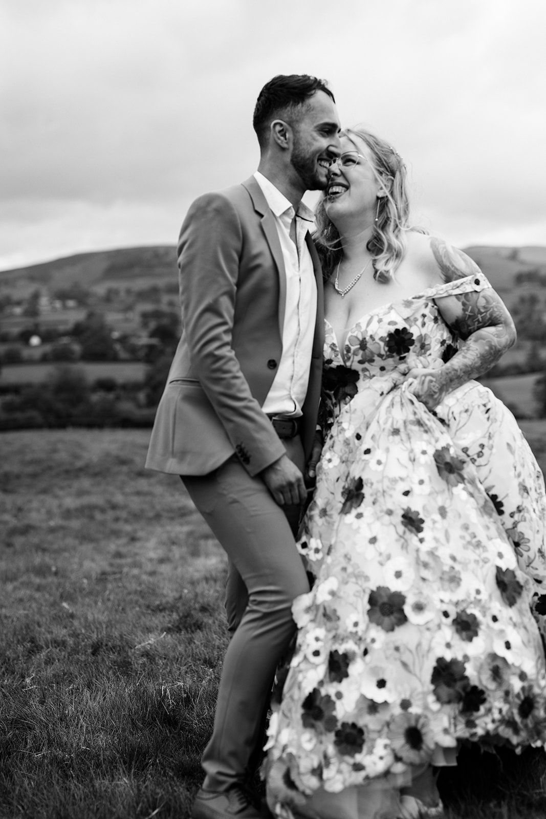 A joyful couple shares a candid moment on their wedding day, with the bride wearing a floral dress against rolling hills.
