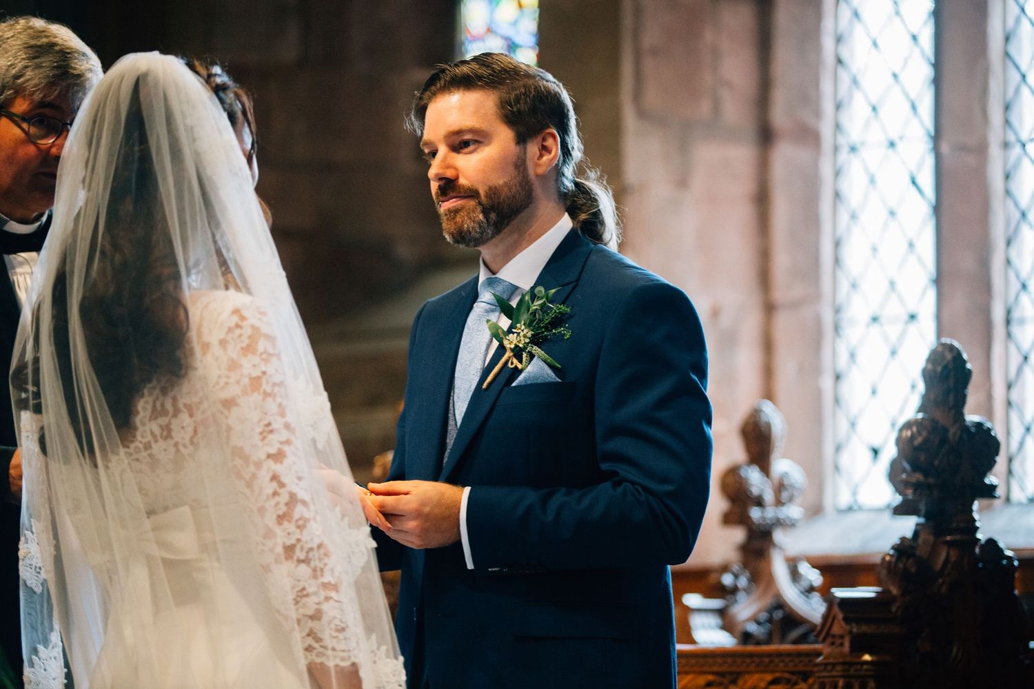 Groom in a navy suit with a boutonniere exchanges vows with a veiled bride during a church wedding ceremony.