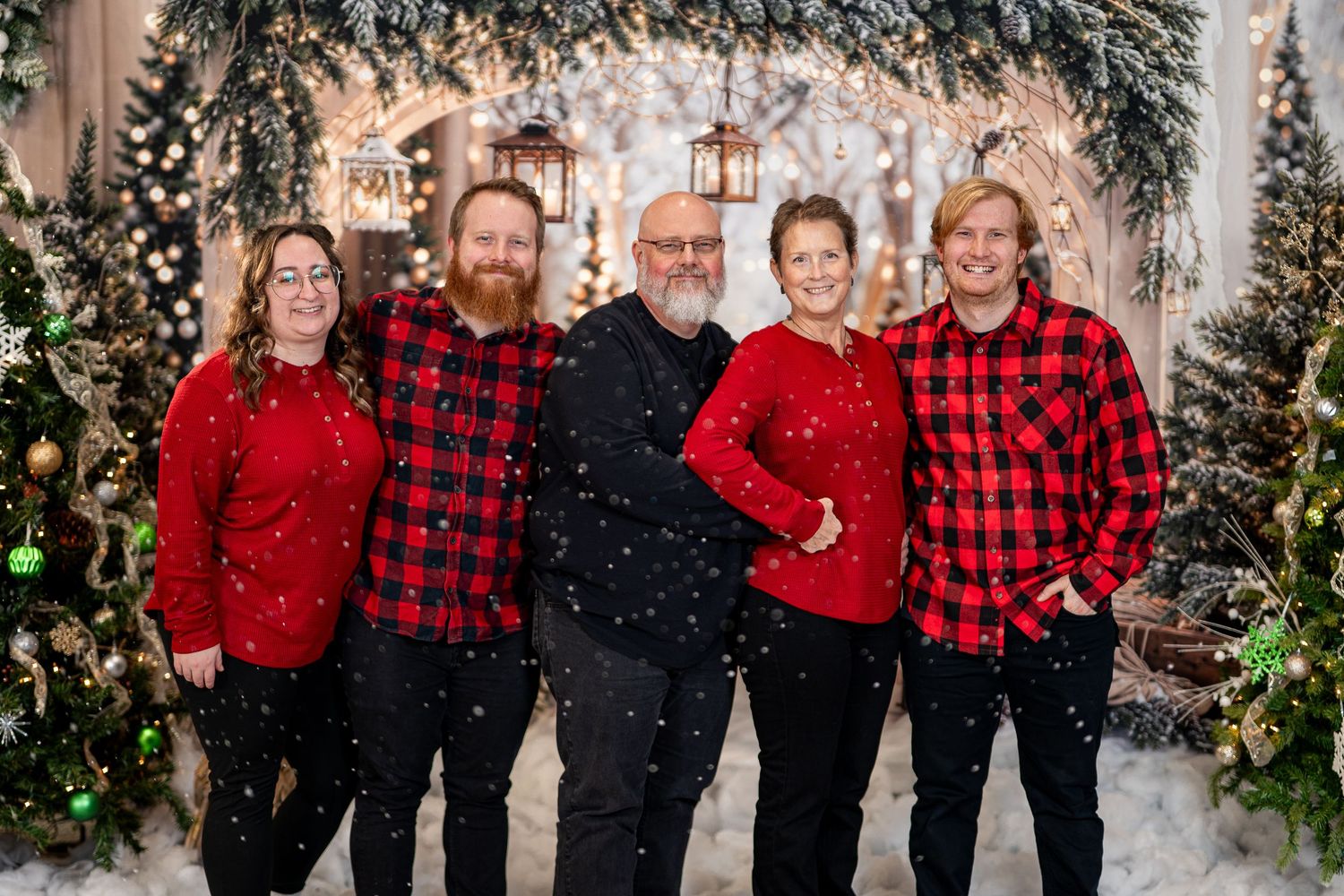 A group wearing coordinated red and black buffalo plaid outfits poses for holiday photos in front of a snowy backdrop.