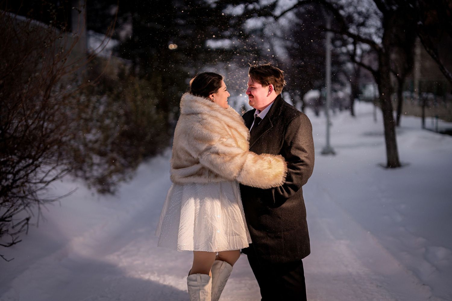 Couple embracing romantically on a snowy winter path, woman in white fur coat and man in dark suit at dusk.