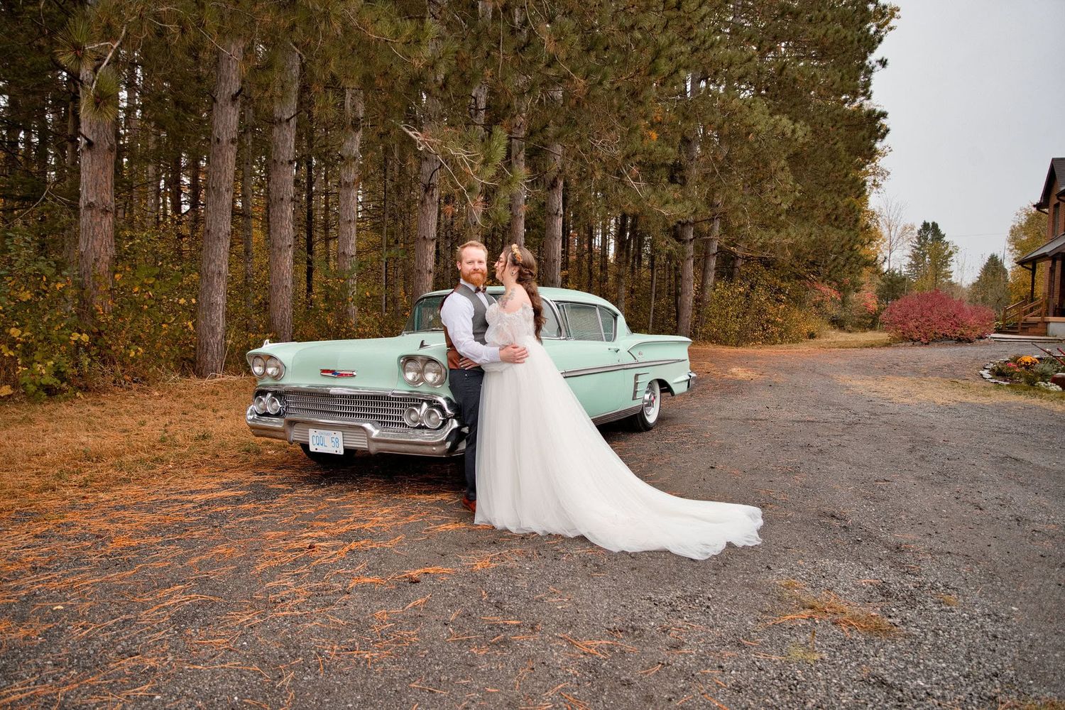 Bride and groom pose beside a vintage mint green 1958 Cadillac surrounded by tall pine trees in autumn.