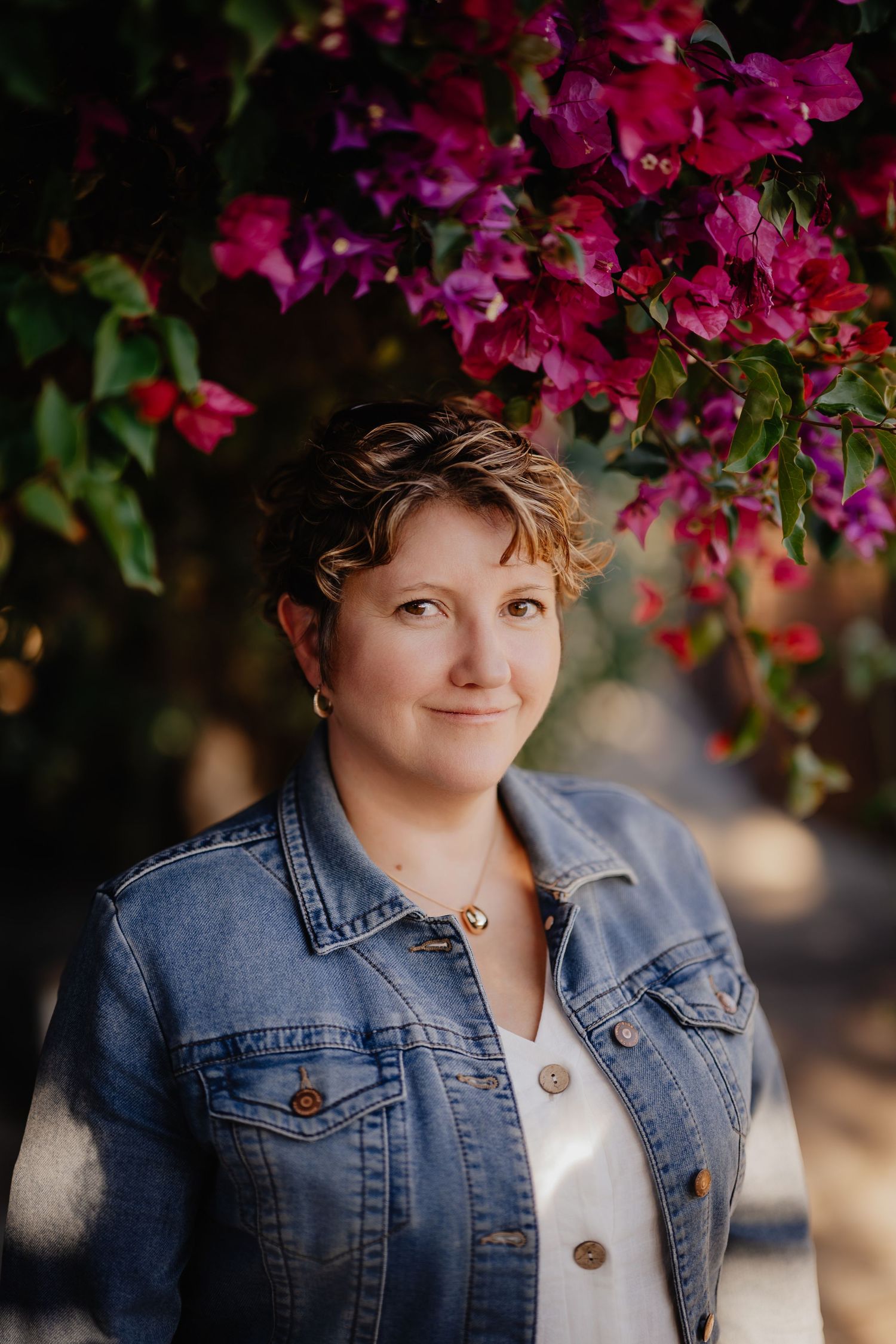 Woman in denim jacket smiling under vibrant pink bougainvillea flowers in a sunny outdoor setting.