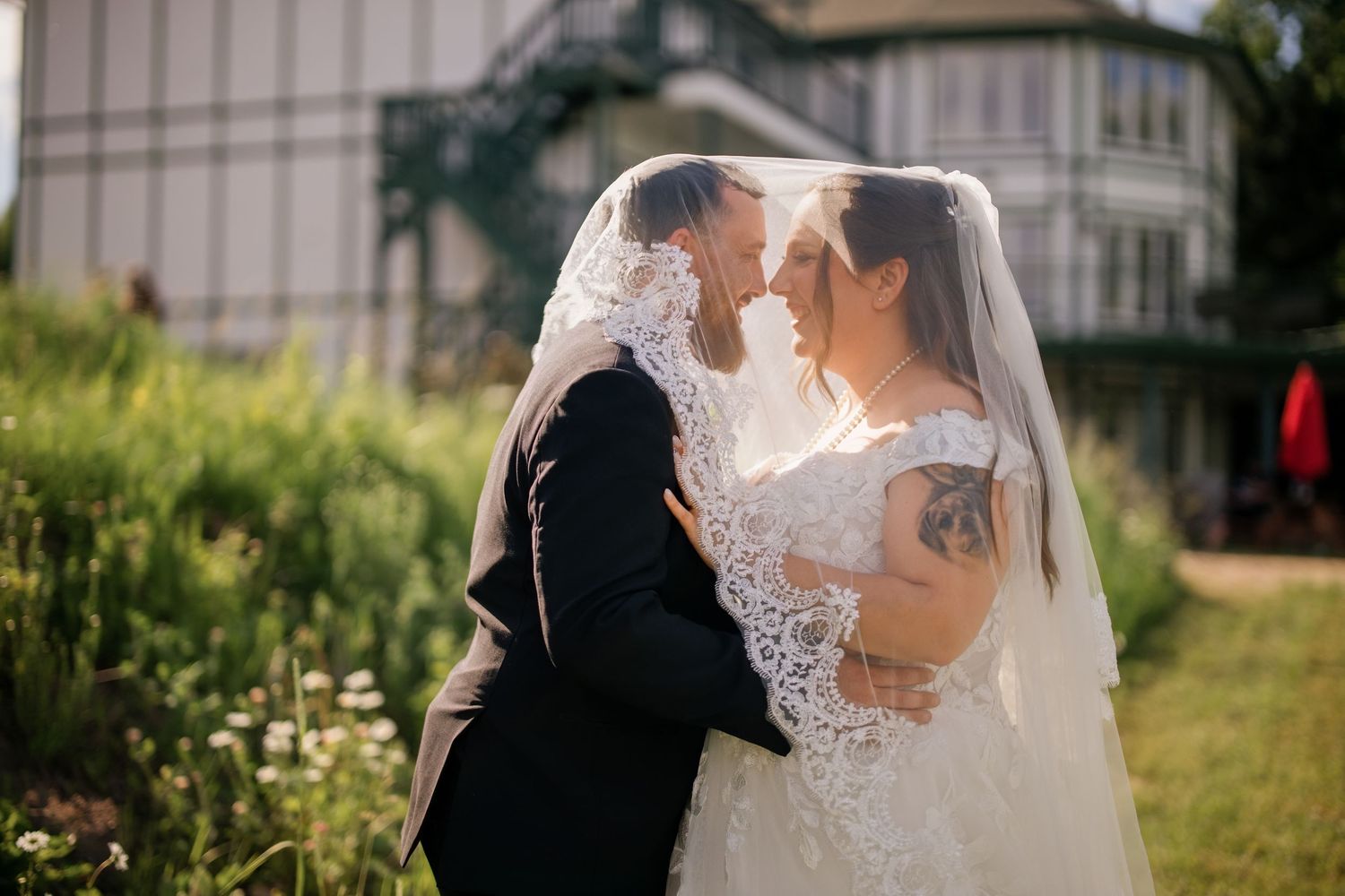 Bride and groom sharing romantic moment under lace veil outdoors at wedding venue surrounded by greenery.