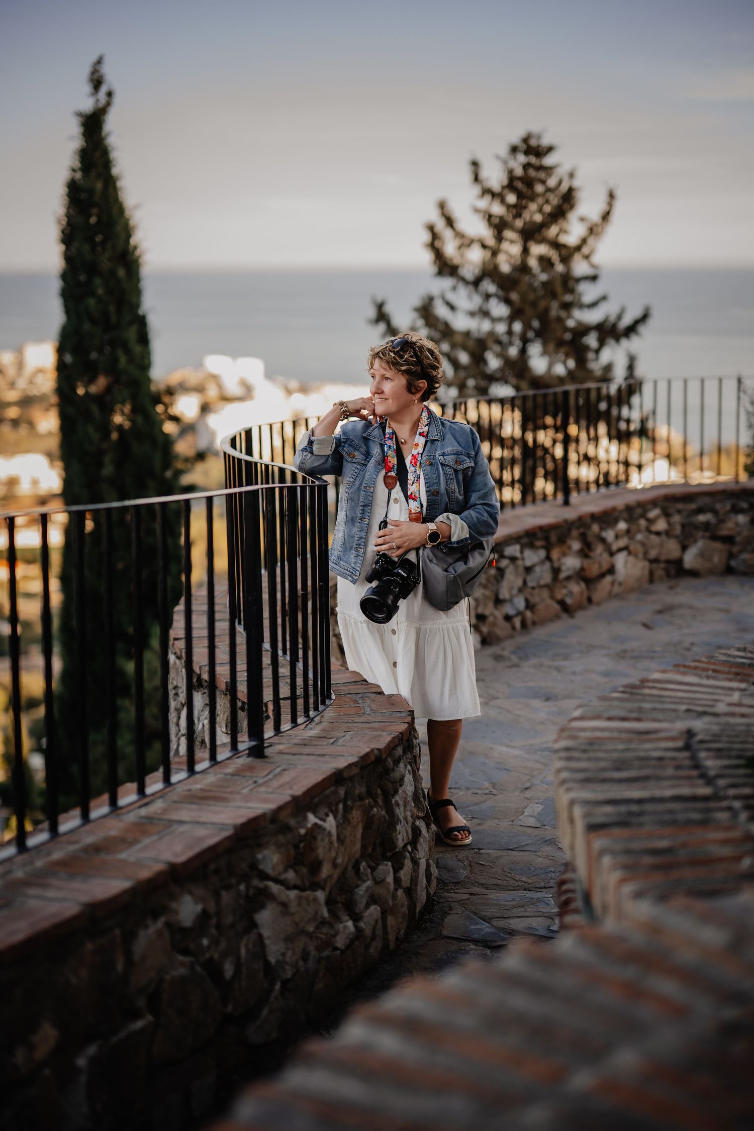 A photographer in denim jacket works at sunset along a stone wall pathway with cypress trees and metal railing.