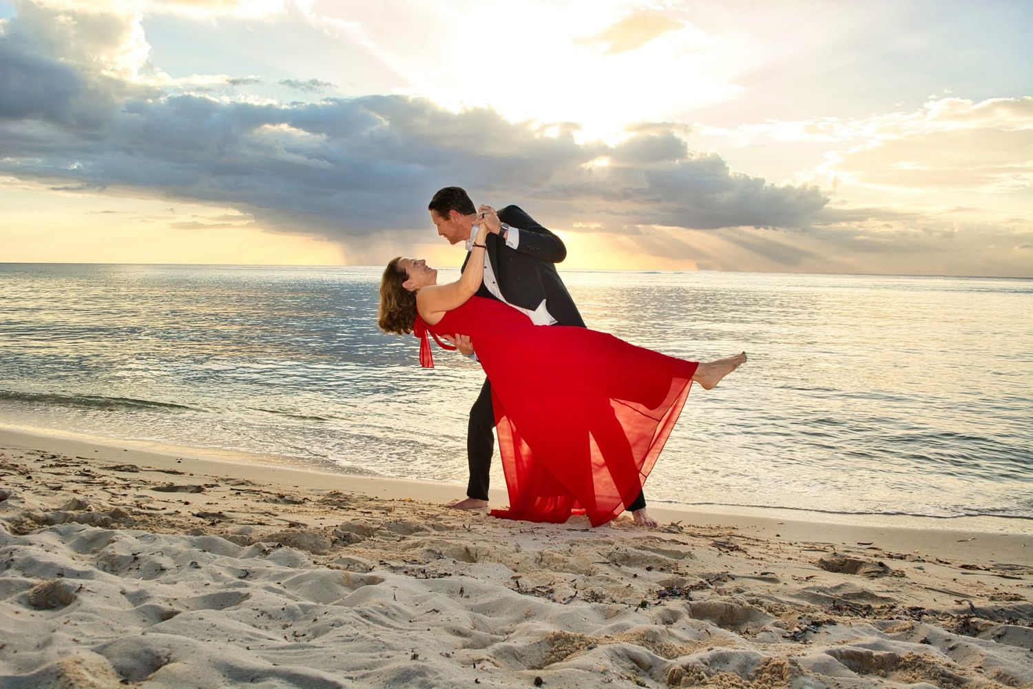 Couple sharing a romantic dip kiss on a sandy beach at sunset, woman in flowing red dress, man in dark suit.