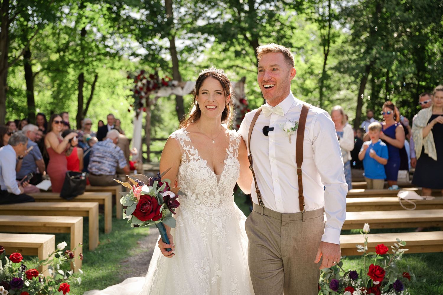 A wedding ceremony takes place outdoors in a wooded setting with wooden benches and red floral decorations.