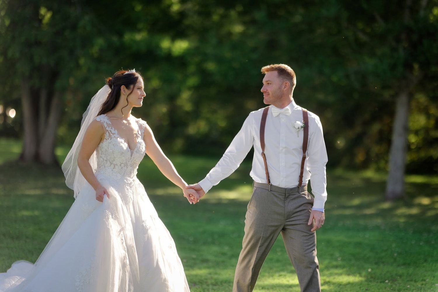 Bride and groom hold hands in a sunlit green park, bride in white gown with veil, groom in suspenders.