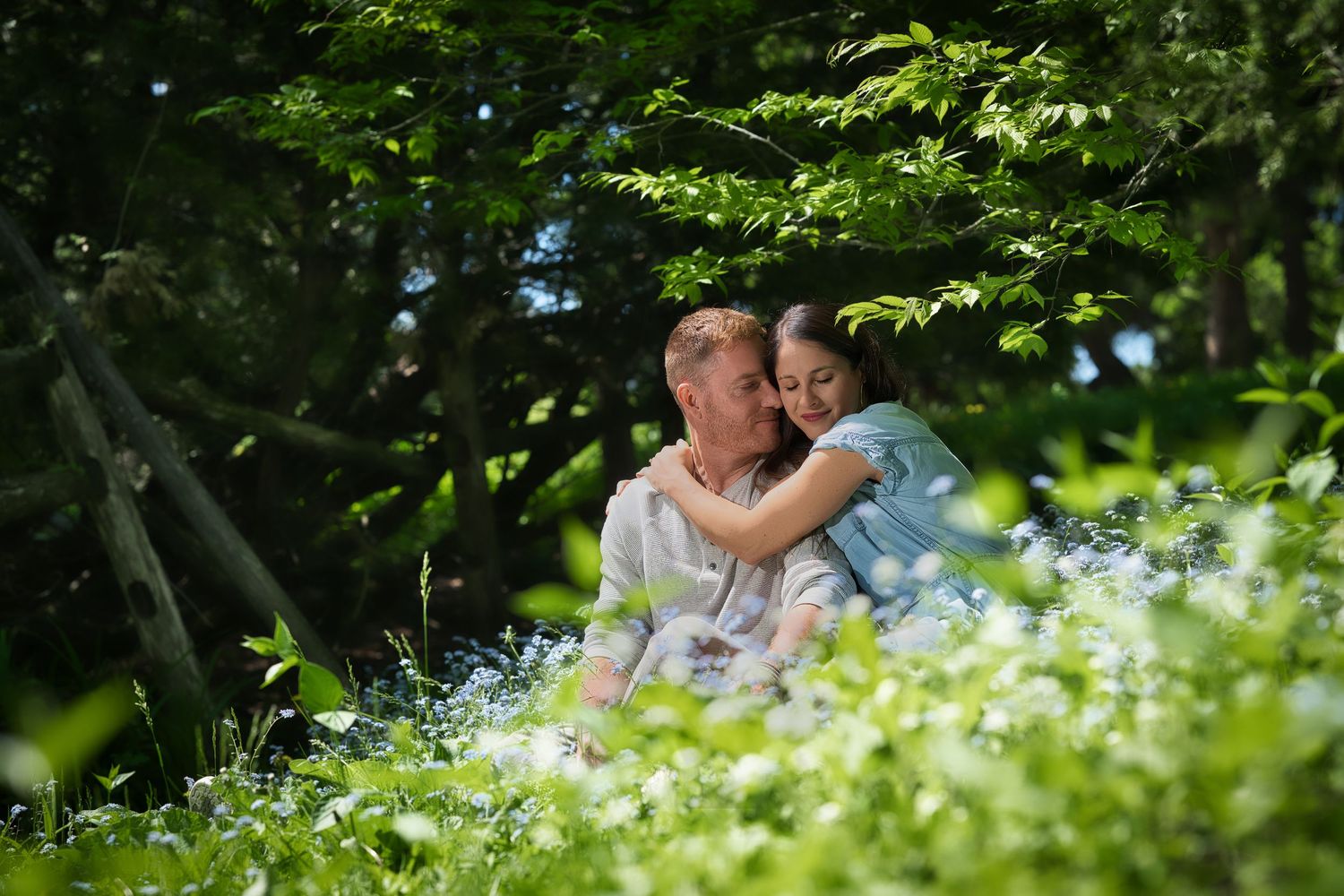 A couple shares a romantic moment surrounded by lush greenery and white wildflowers in a natural outdoor setting.