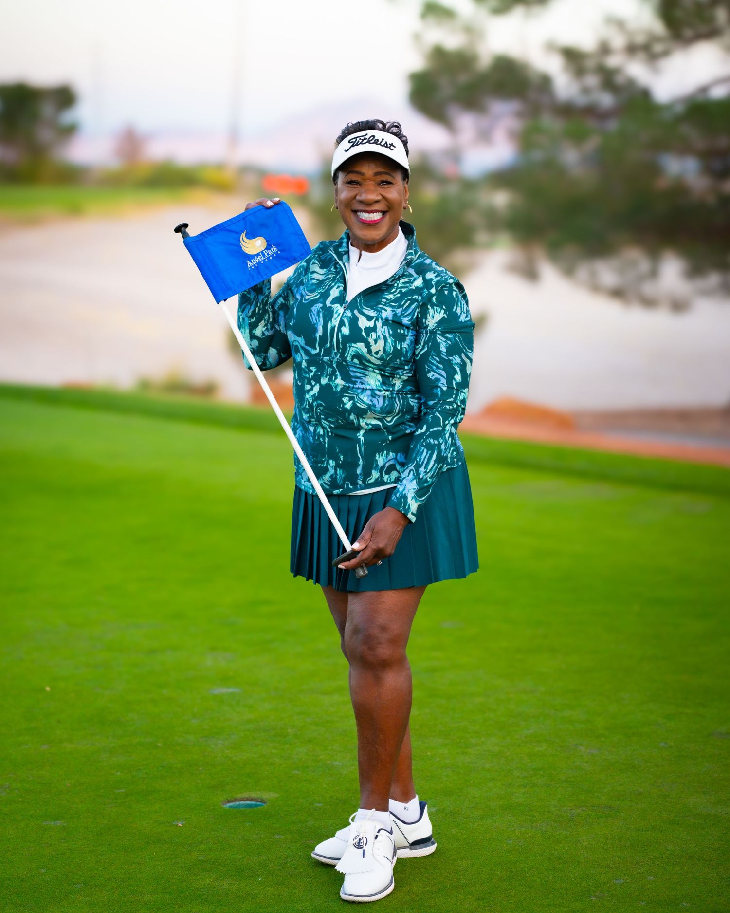 Smiling woman in teal golf outfit holds blue flag on golf course green.