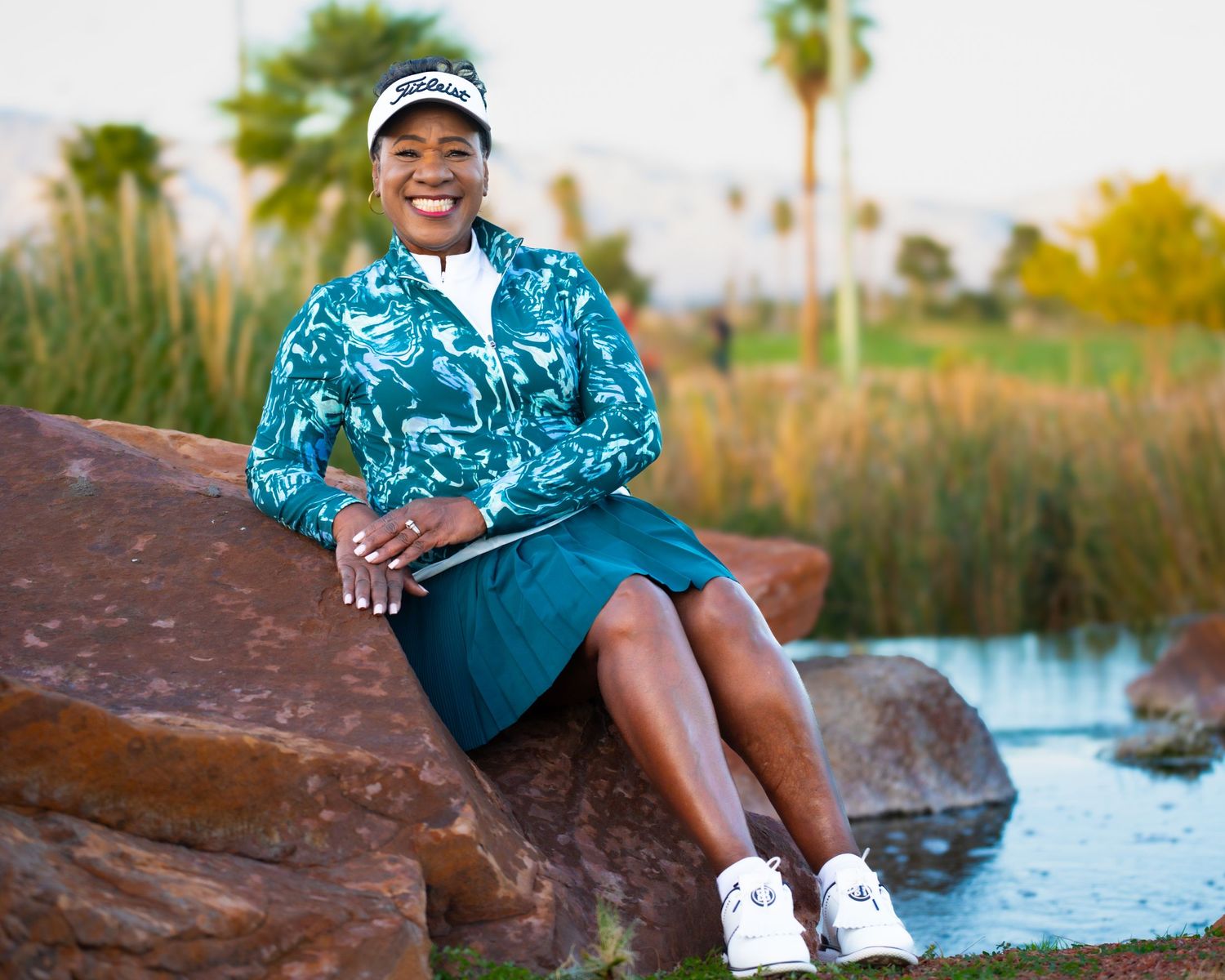 Woman in teal golf outfit sits relaxed on rocks near water at golf course.