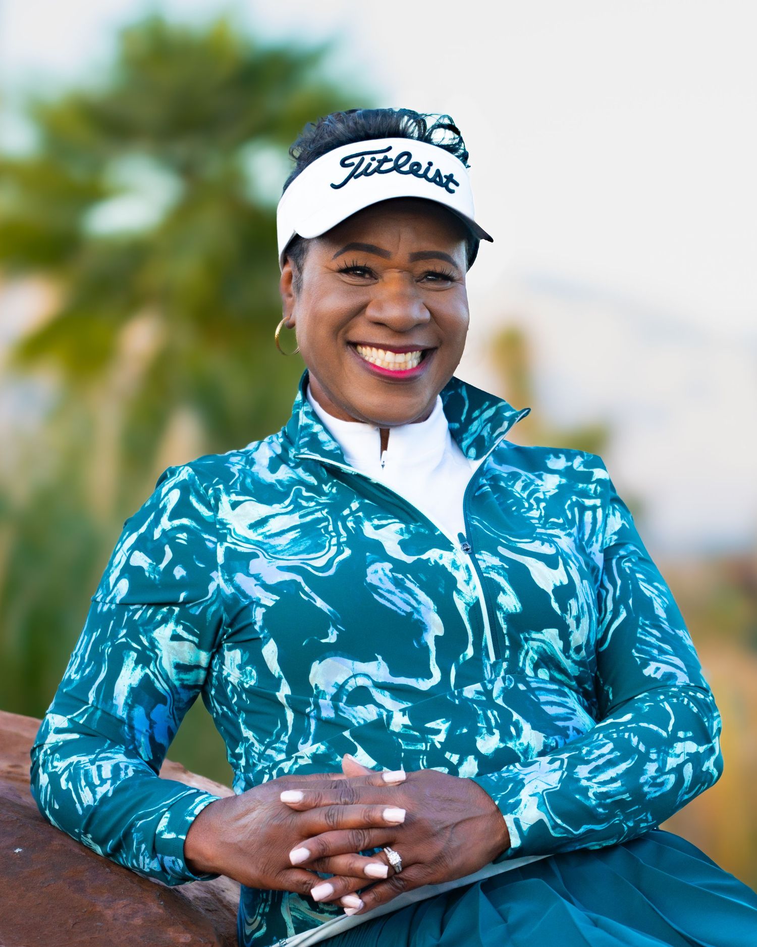 Smiling woman wearing Titleist visor and teal golf jacket poses outdoors near palm trees.