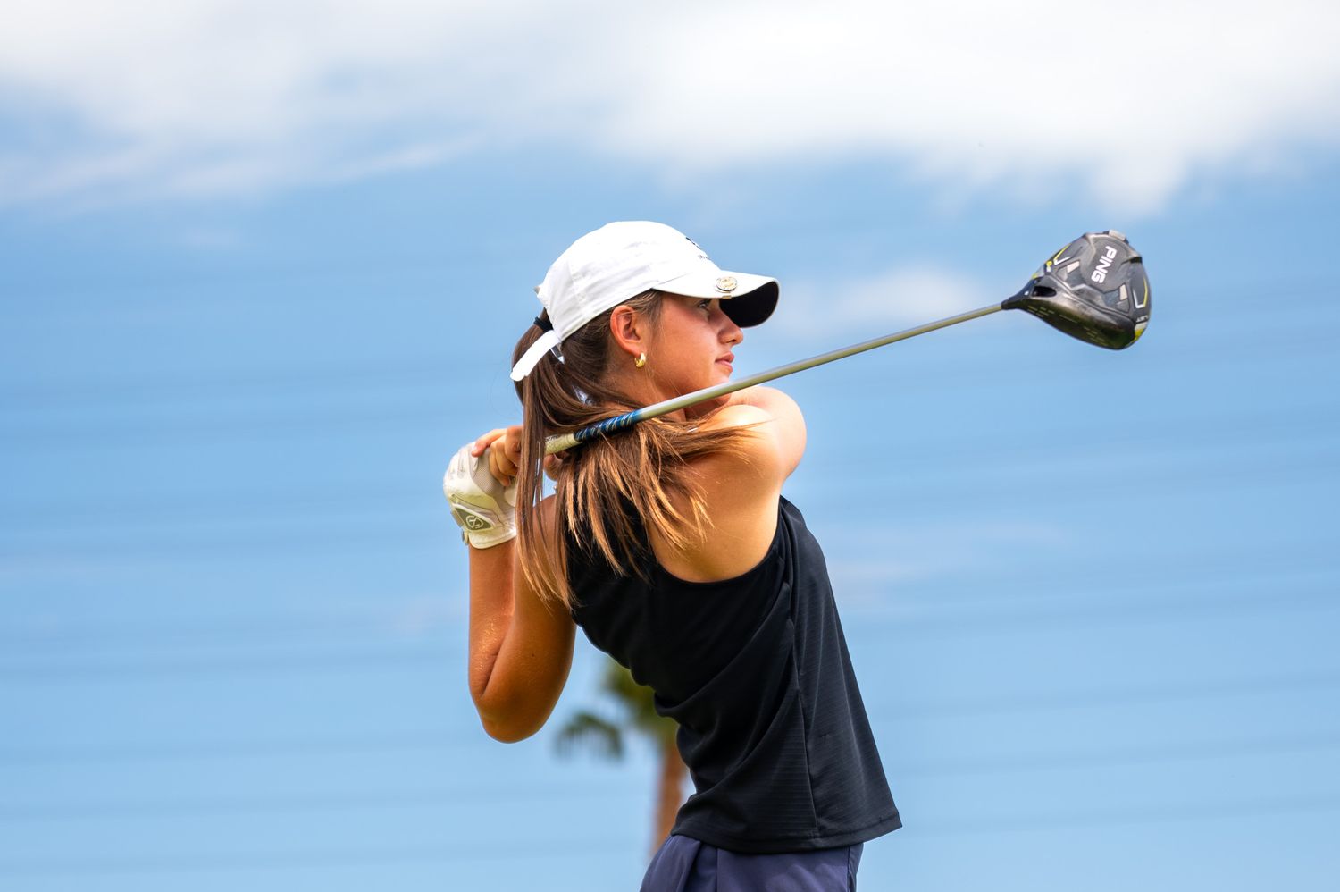 Female golfer in black sleeveless top and white cap completing a powerful golf swing against a blue sky.