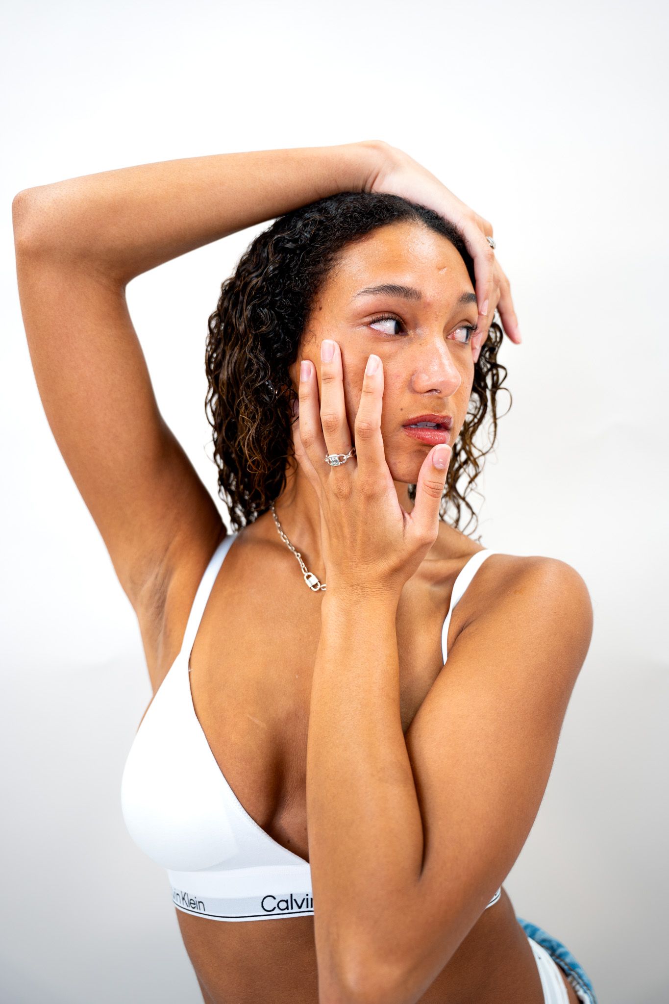 Young woman in white Calvin Klein bralette posing with hands on face and arm raised in white studio.