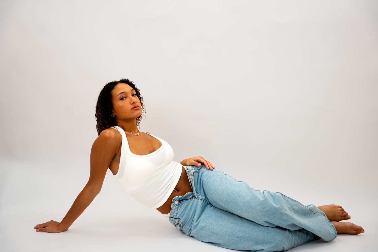 Young woman in white tank top and jeans reclining gracefully on white studio floor.