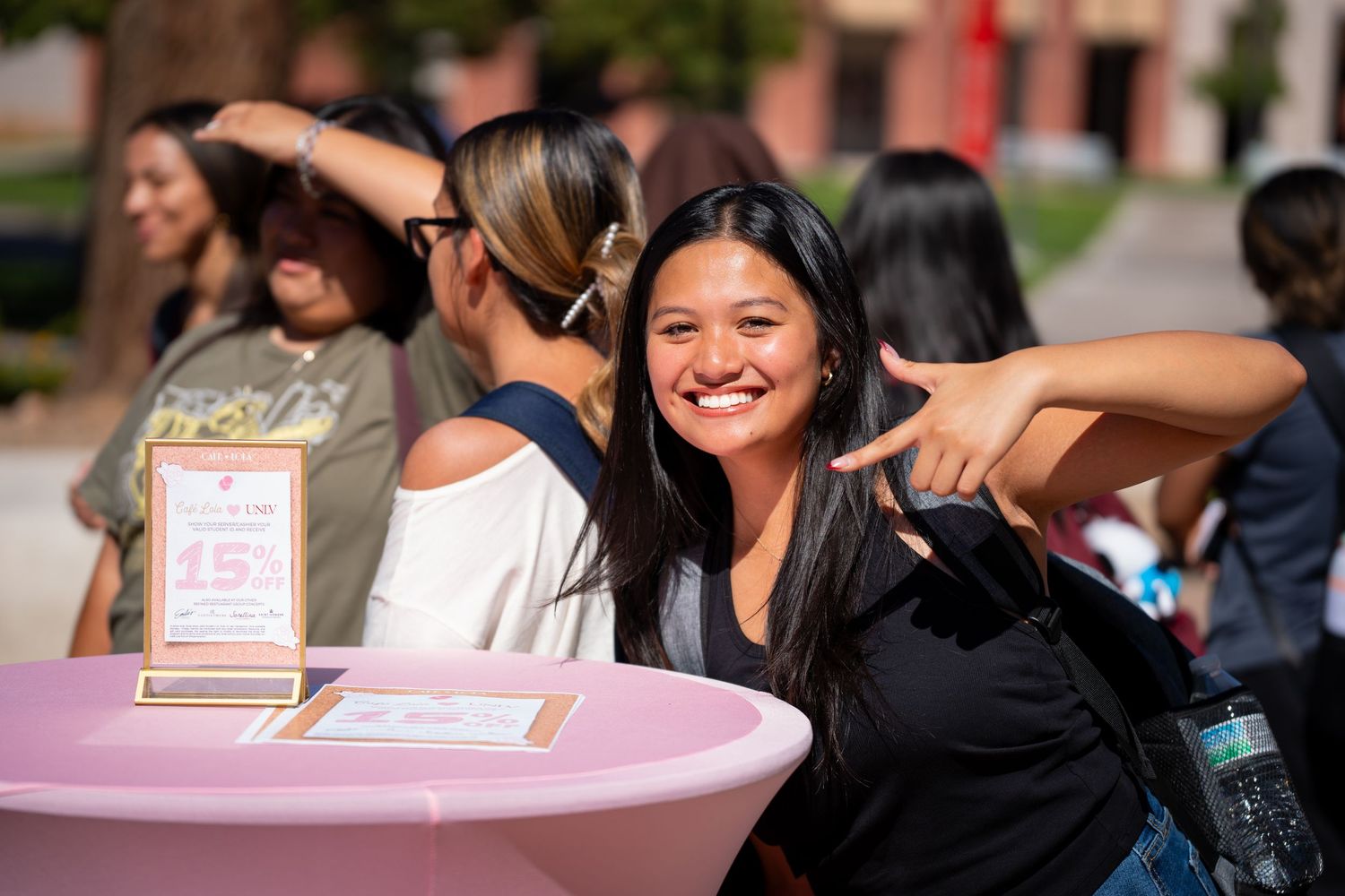 Smiling young woman points finger gun at camera while standing at a pink table at outdoor event.