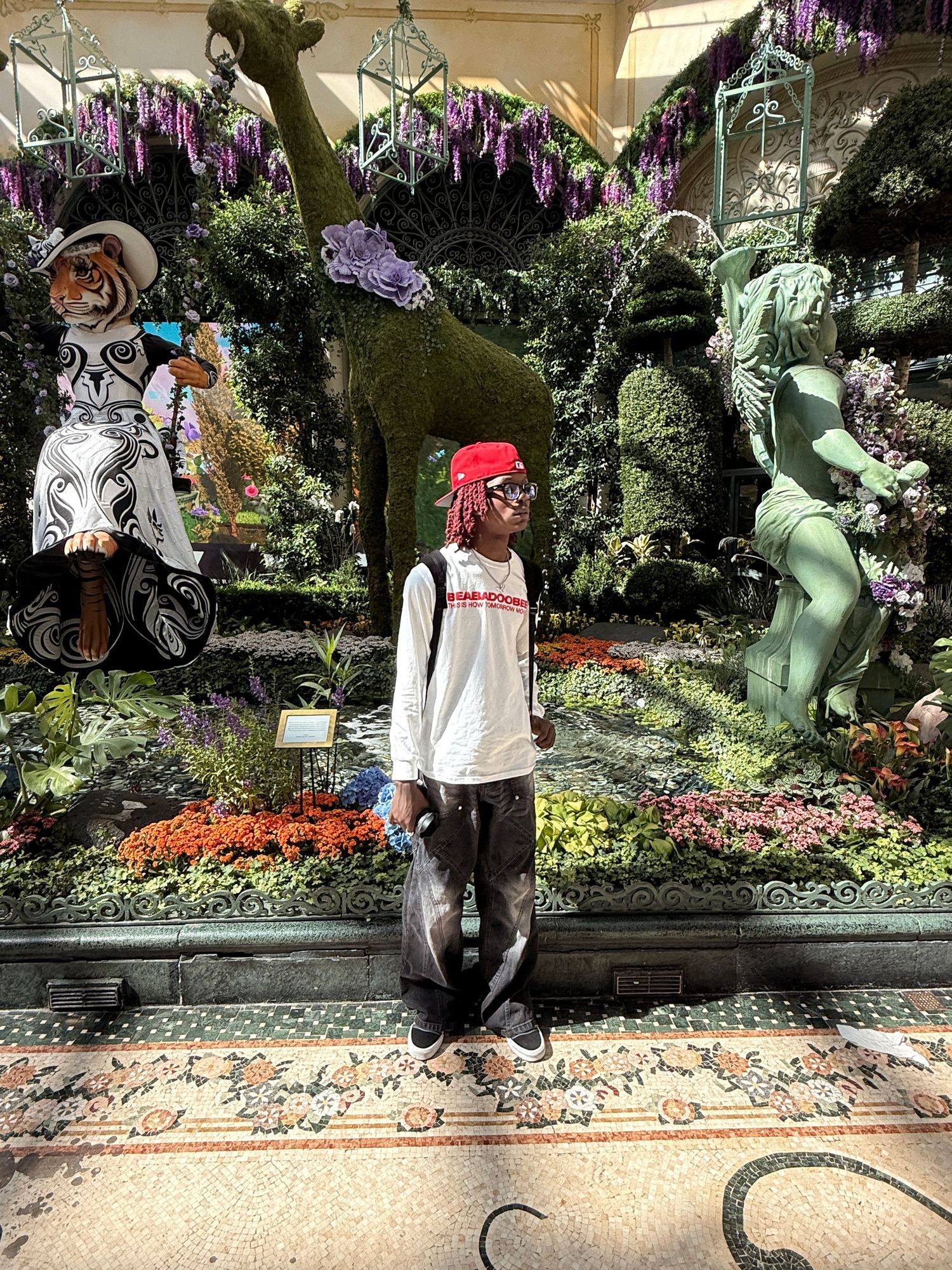 Young man in red hat and white long sleeve standing before elaborate floral garden display.