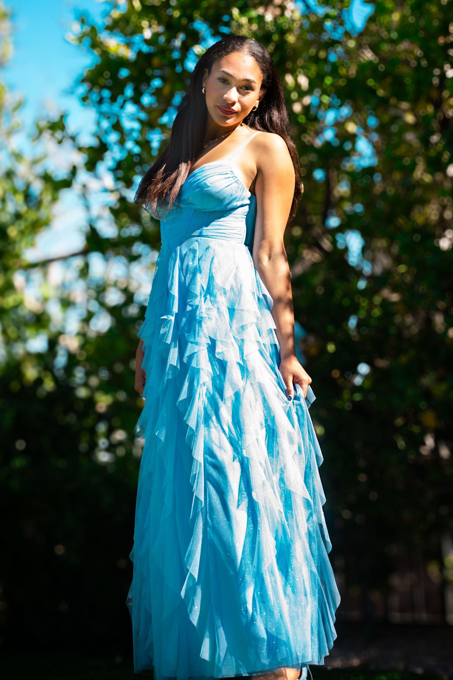 Young woman in a light blue strapless ruffled dress poses outdoors with green trees in background.