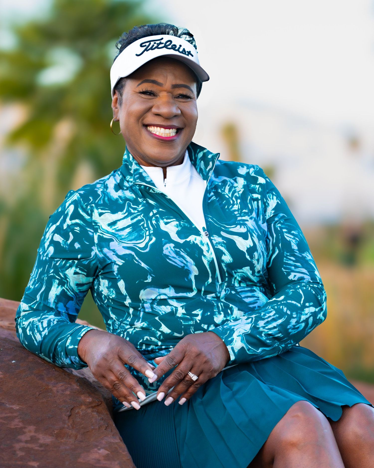 Cheerful woman in teal patterned golf jacket and Titleist visor smiles while seated on rocks.