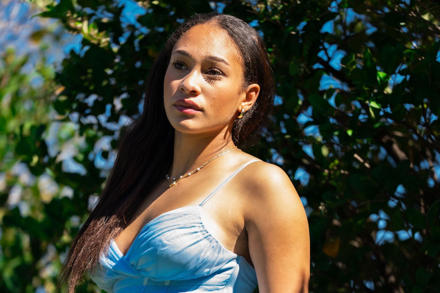 Close-up portrait of a young woman in a light blue dress standing near lush green leaves outdoors.