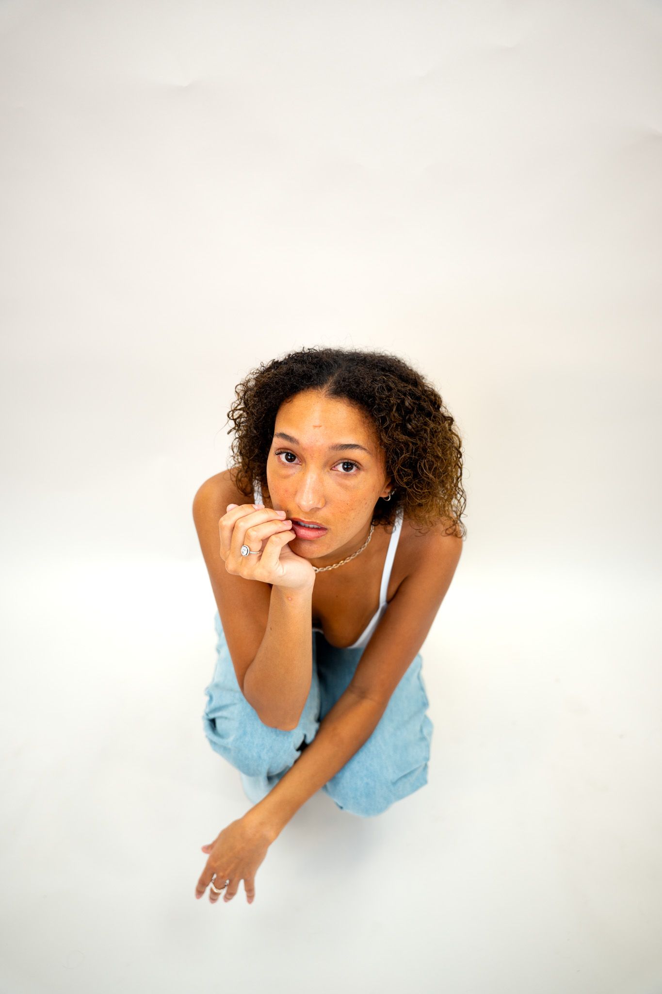 Young woman with curly hair in white crop top and jeans sitting on floor looking up at camera.