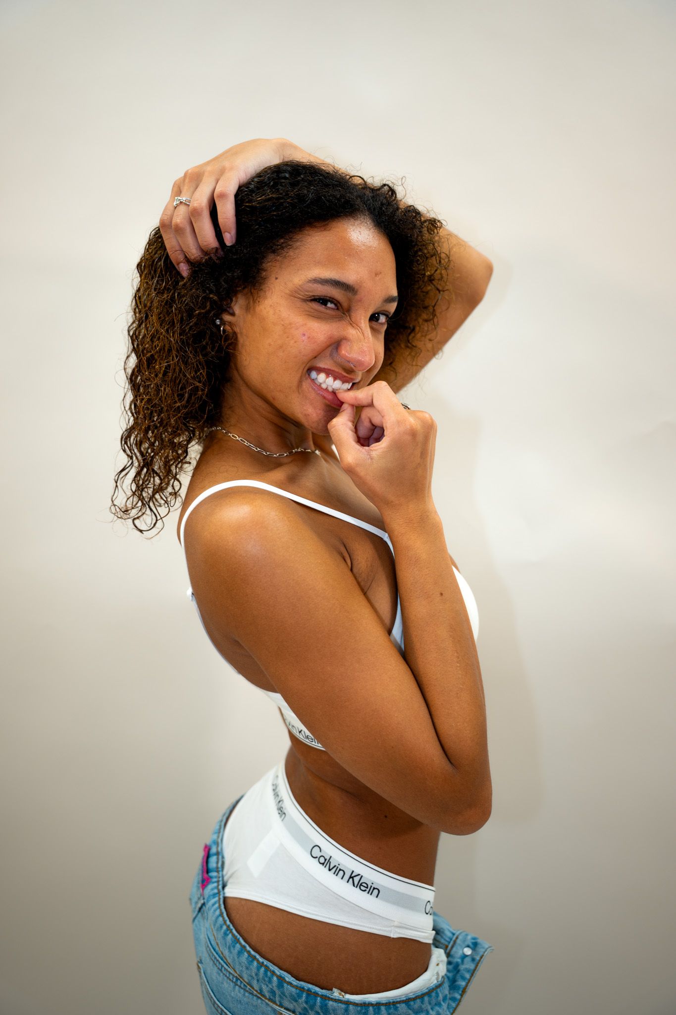 Smiling young woman in Calvin Klein bralette and jeans poses playfully against white background.