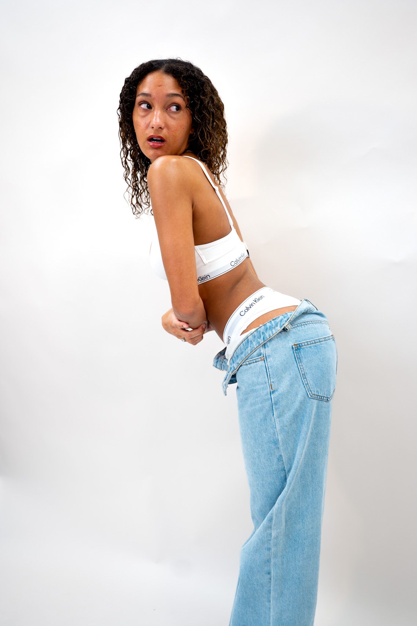 Young woman in white bralette and low-rise jeans looking back over shoulder in white photo studio.
