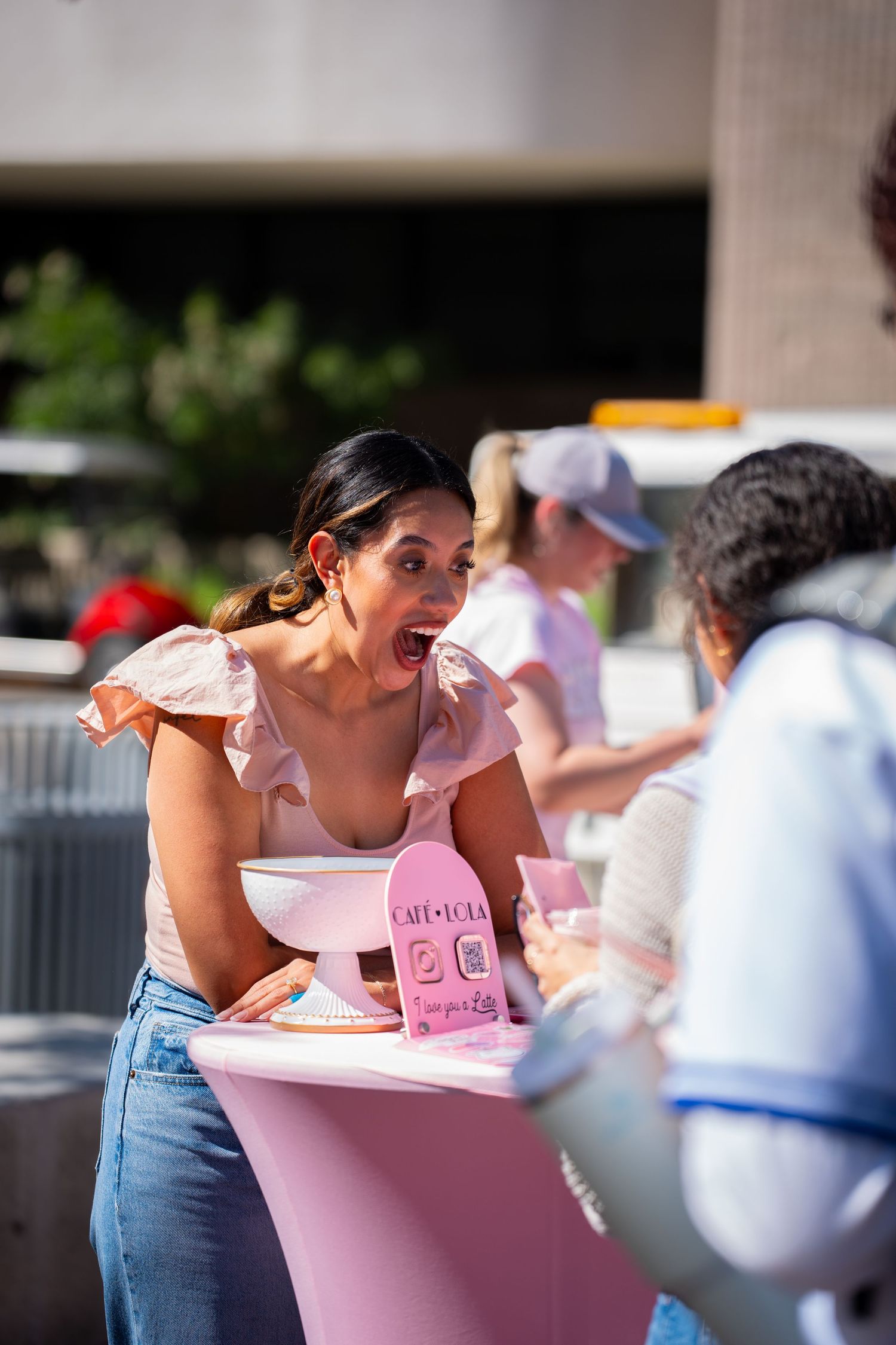 Excited woman with open mouth expression leaning over a pink Cafe Lola booth table at an outdoor event.