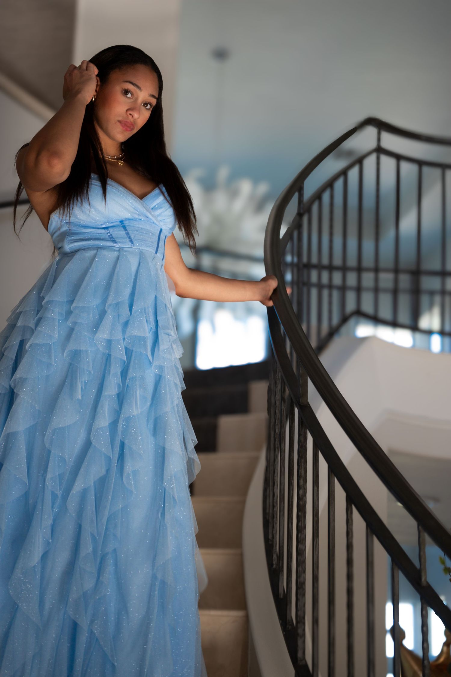 Young woman in a blue ruffled gown poses on an elegant staircase with black iron railing.