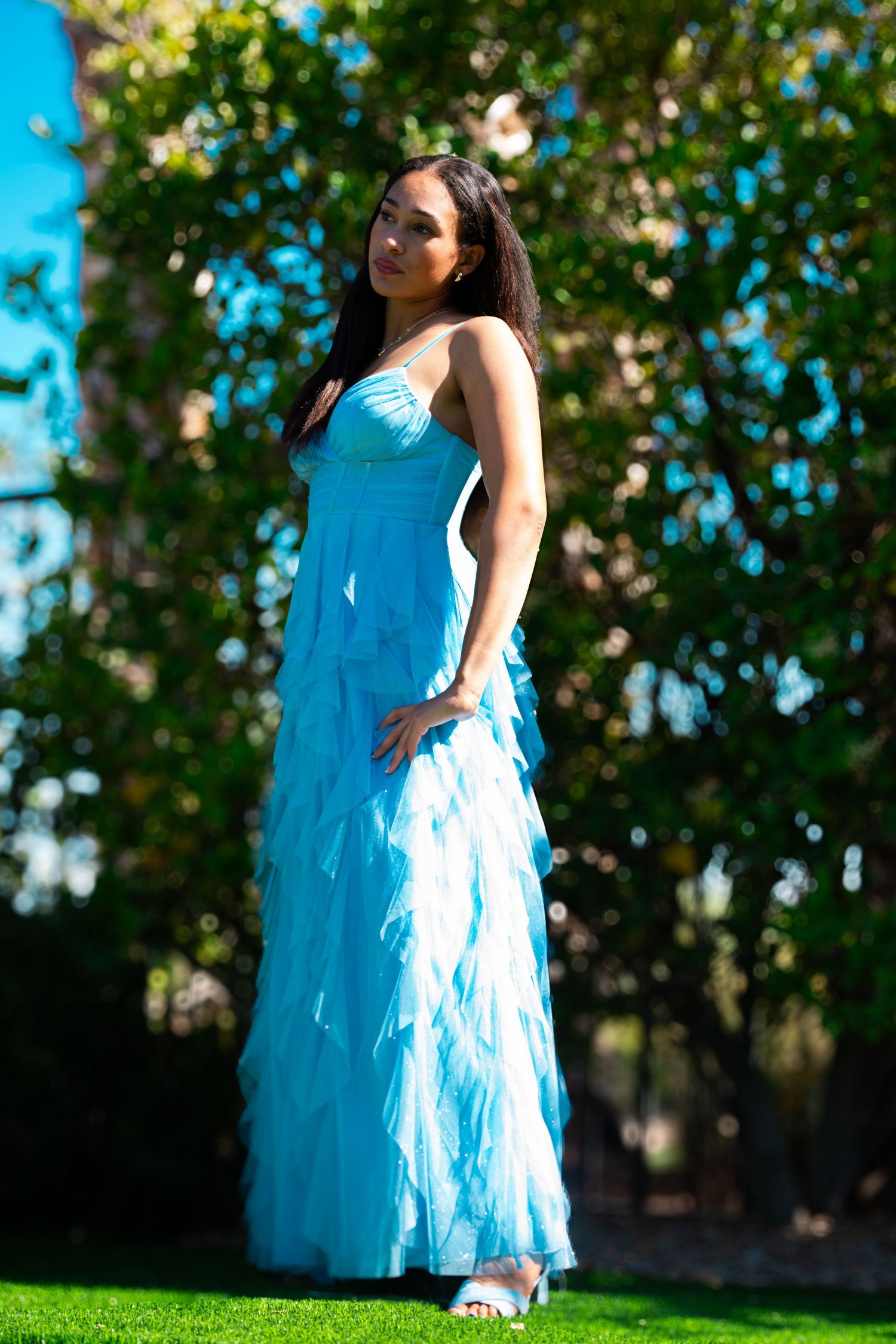 Young woman in elegant light blue ruffled formal gown posing outdoors on green grass with lush trees in background.
