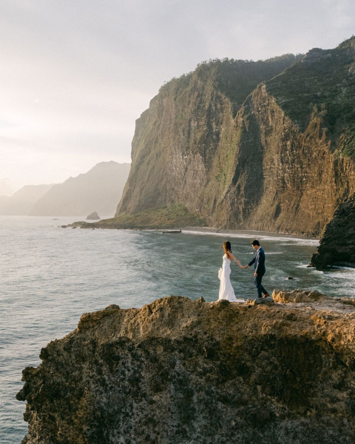 Couple in wedding attire holding hands on a rocky cliff overlooking the ocean with dramatic green cliffs behind.