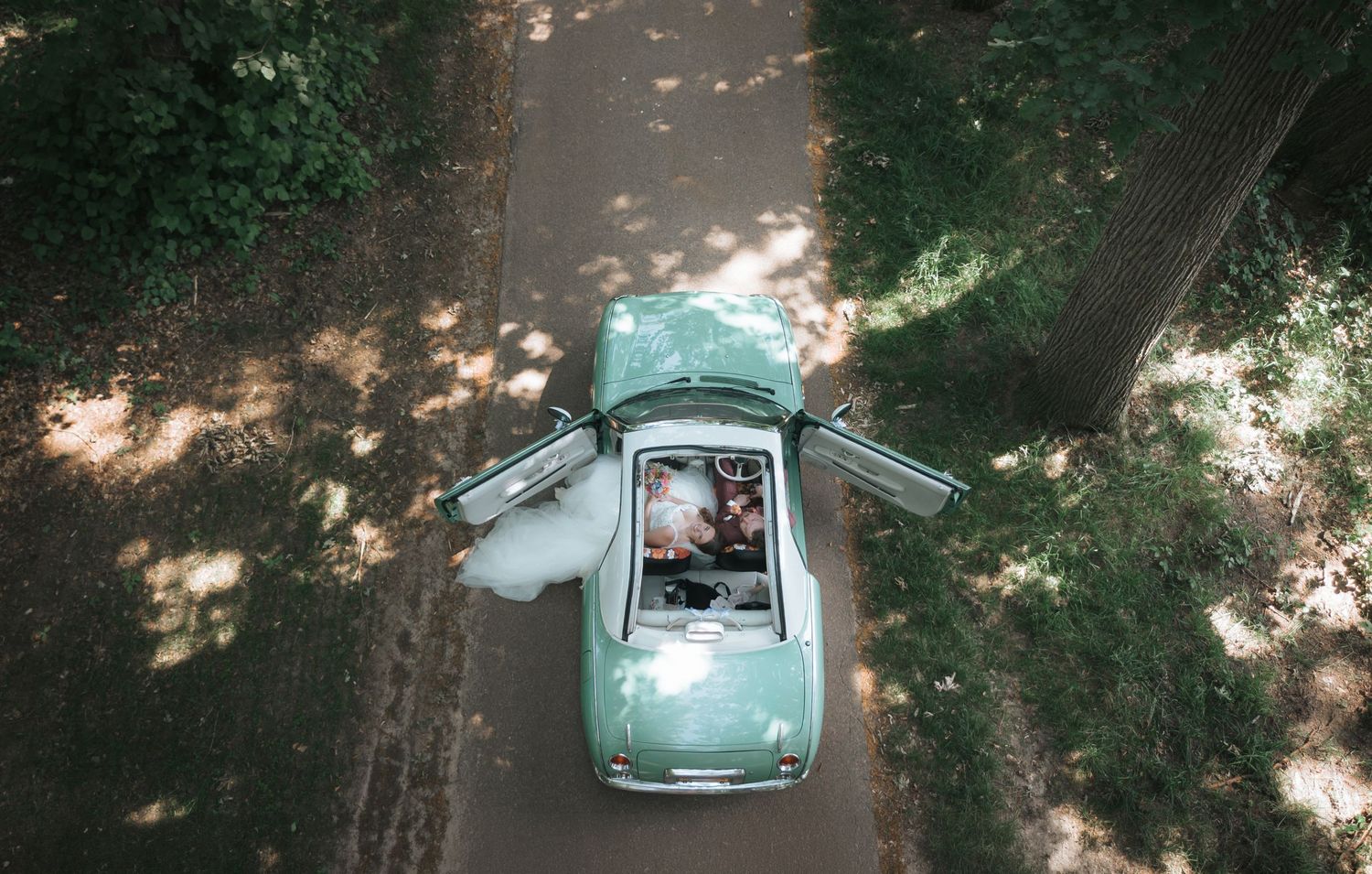 Aerial view of a mint green vintage convertible car driving through a tree-lined path with newlyweds.