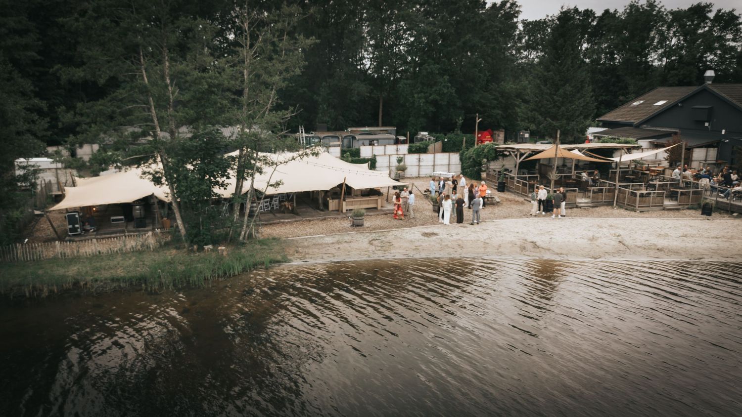 Lakeside campsite with tents, canopies, and visitors relaxing on sandy shore surrounded by lush green trees.