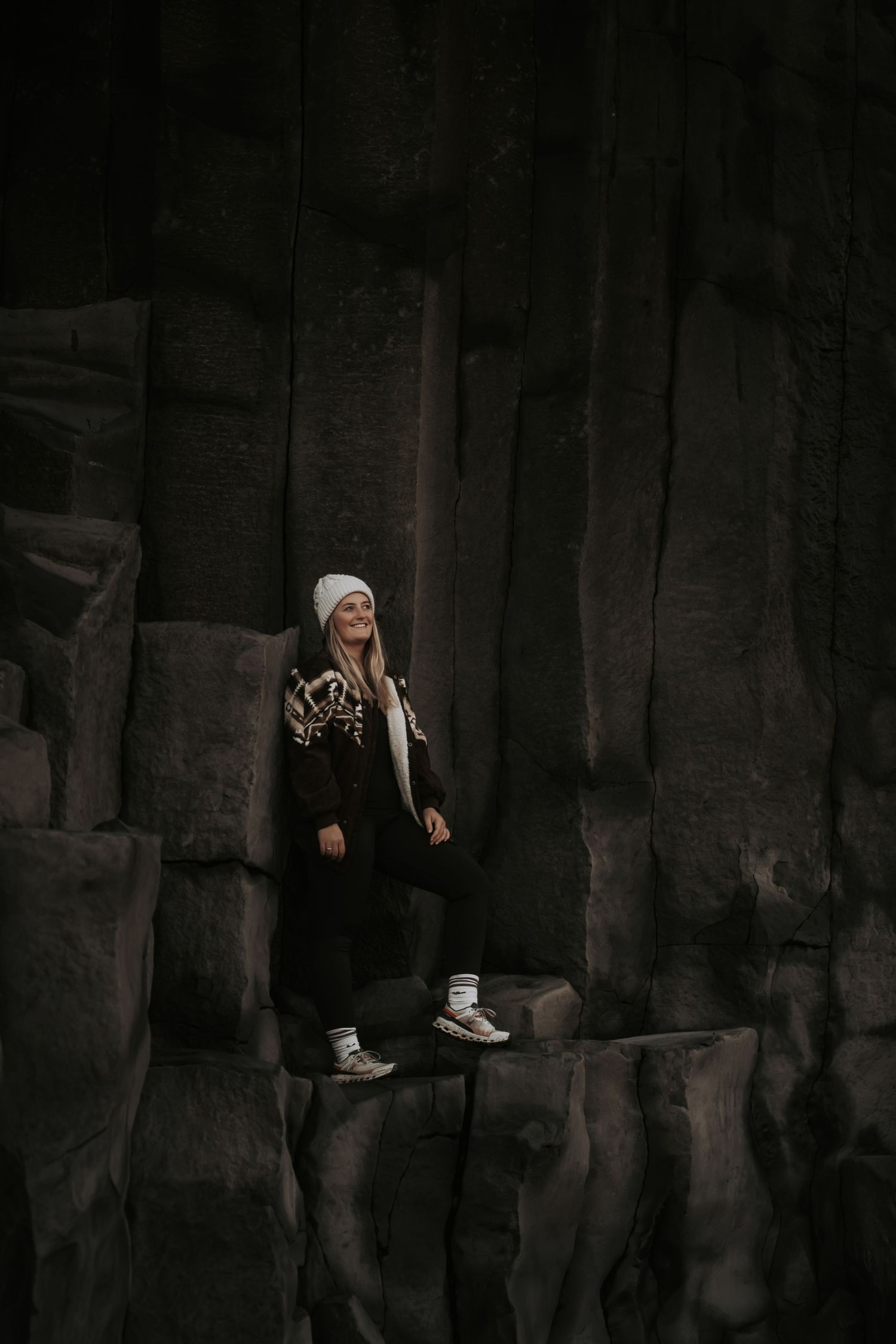 Woman in winter clothes sitting on dark basalt rock columns in a dramatic landscape.