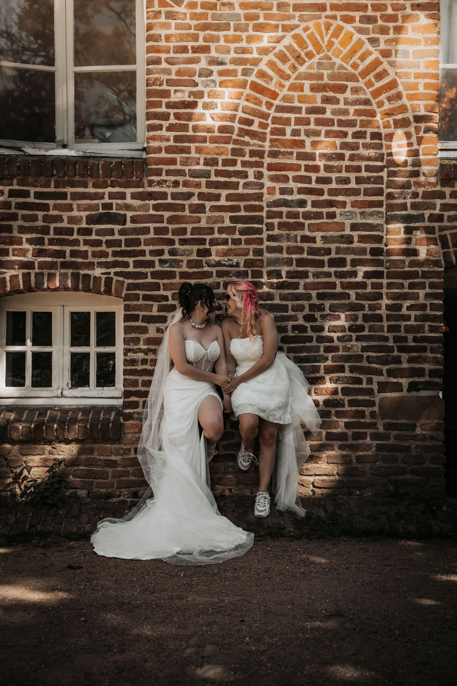 Two brides in white wedding gowns sit closely together against a rustic red brick wall.