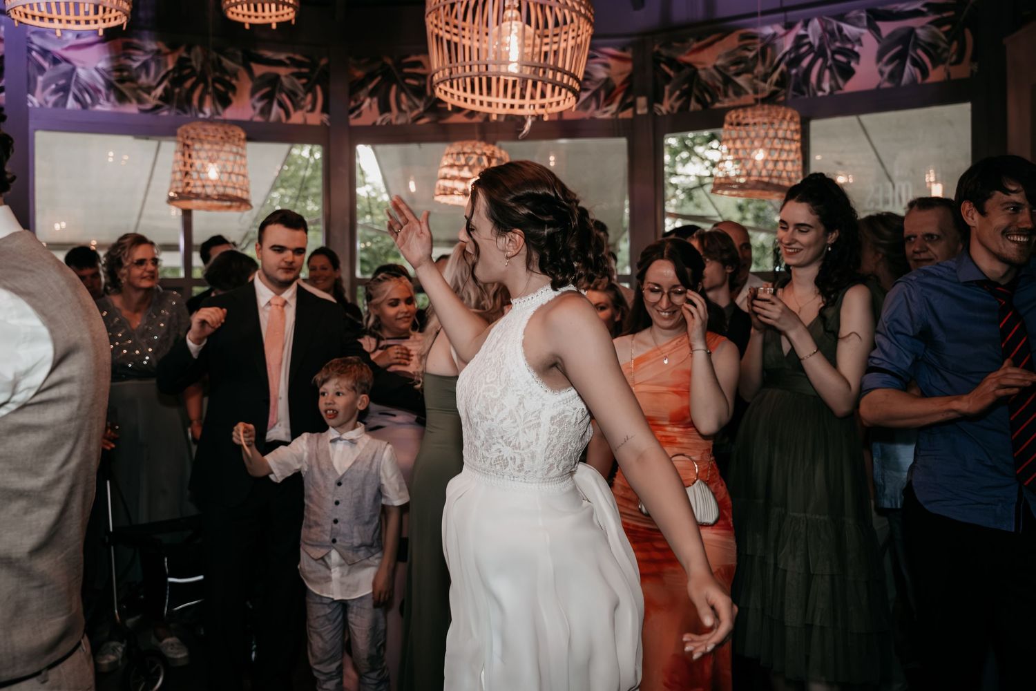 Bride in lace wedding gown dancing joyfully with guests including young boy at reception.