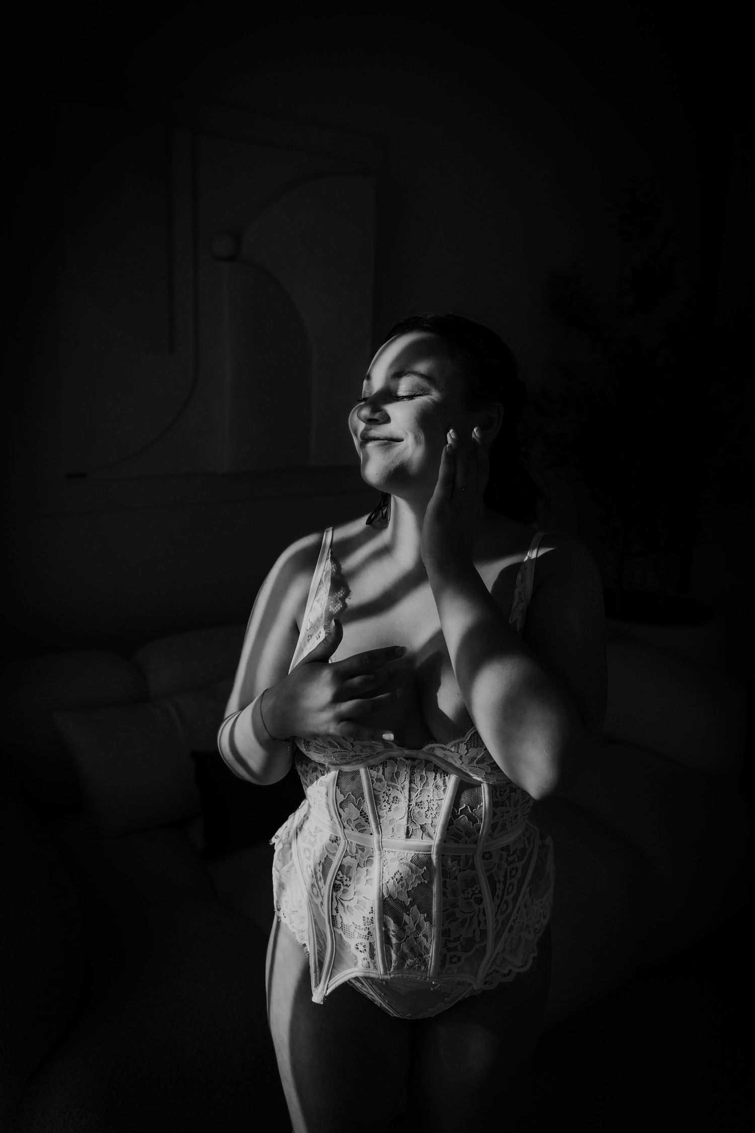 Black and white boudoir photo of woman in lace corset smiling with eyes closed in moody light.