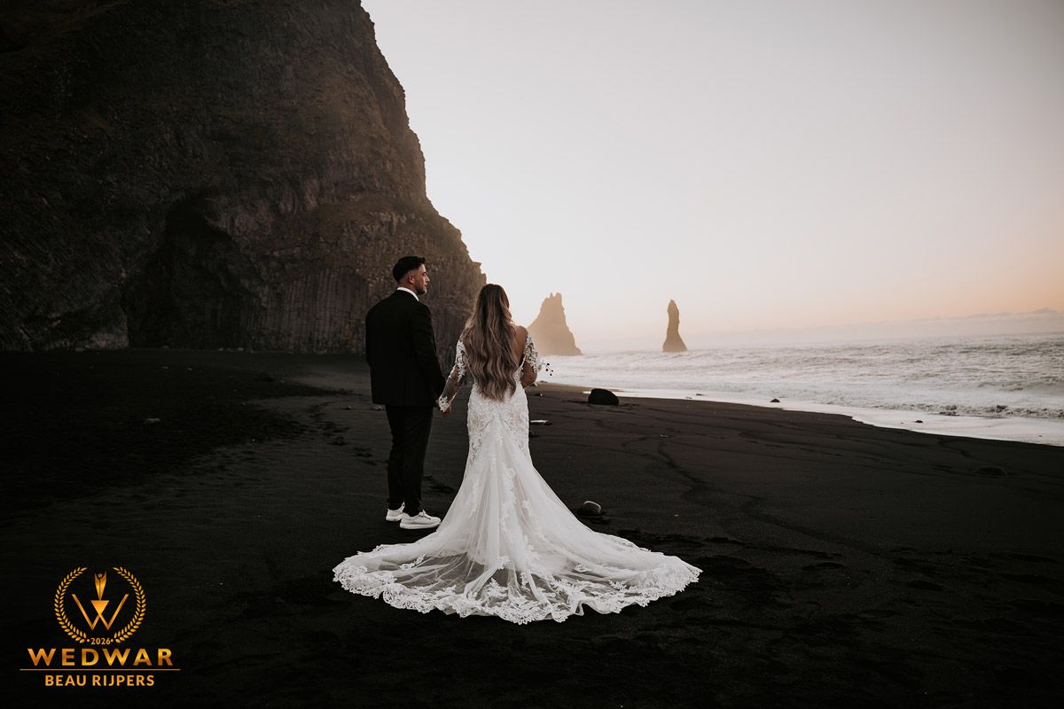 Couple in wedding attire standing on black sand beach in Iceland with dramatic sea stacks and cliffs at sunset.