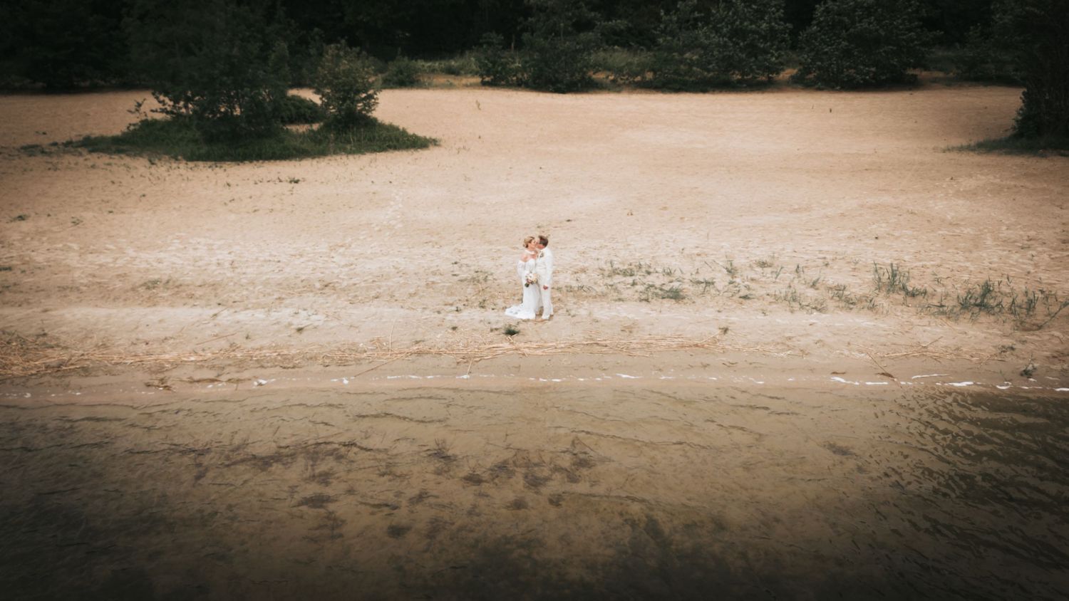 Bride in white wedding dress walking alone along a sandy beach near the water's edge.