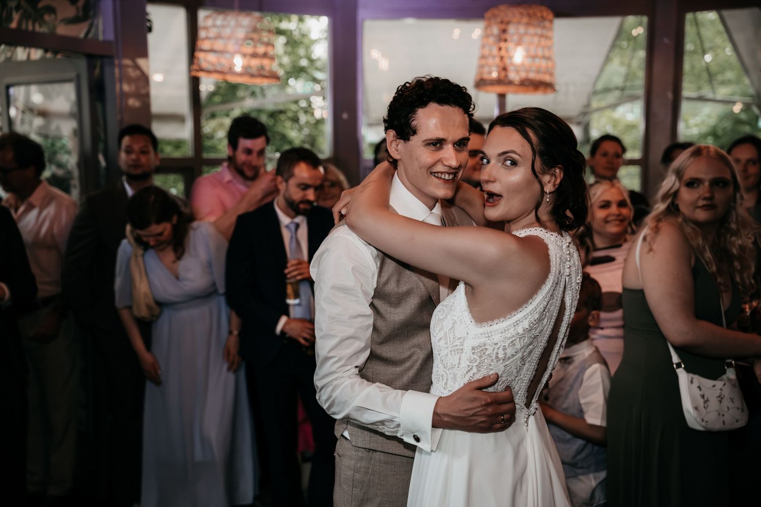 Bride and groom sharing a joyful first dance embrace surrounded by wedding guests at indoor reception.