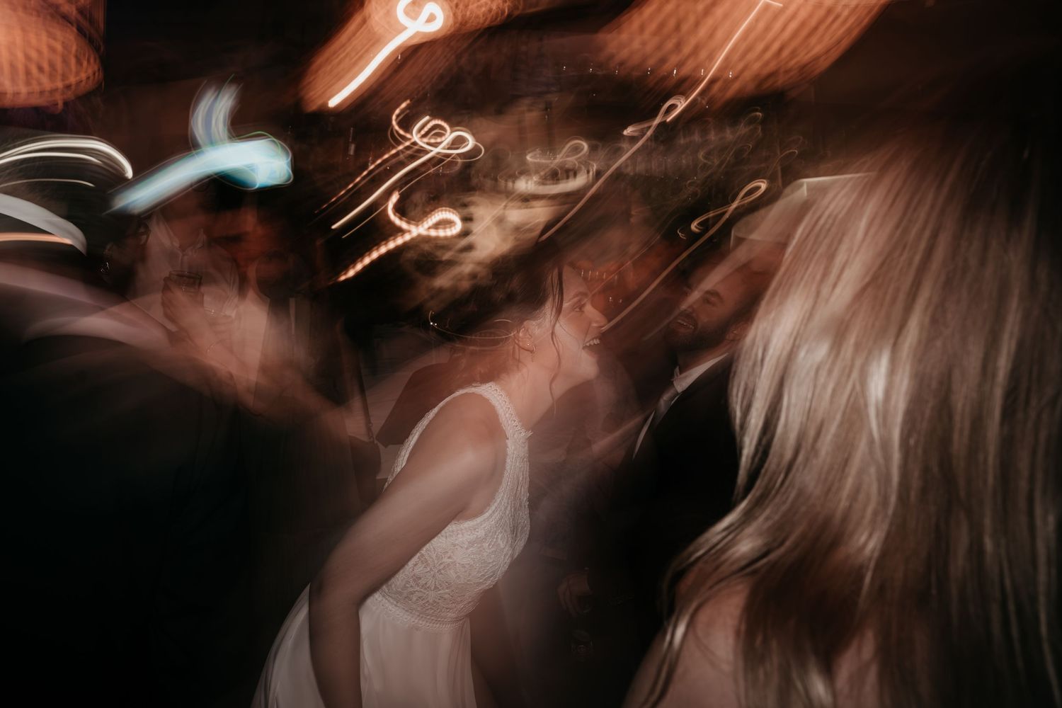 Artistic motion blur of bride dancing with guests under glowing neon lights at wedding reception.