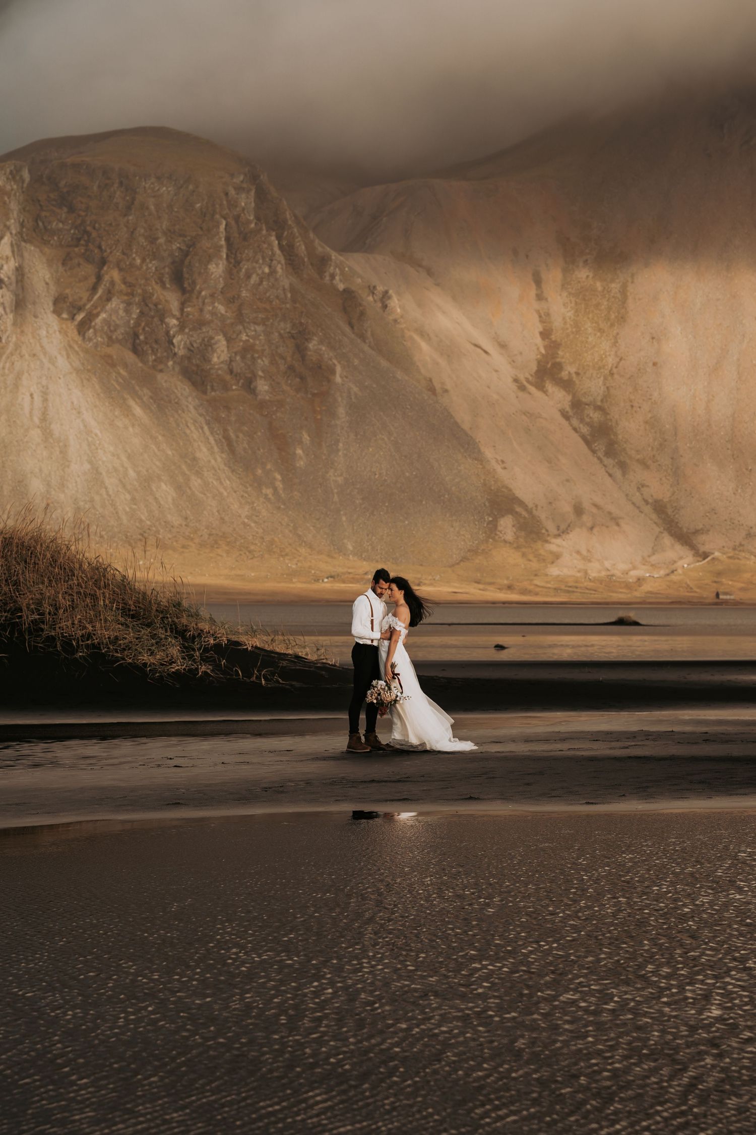 Bride and groom embracing on a dark sandy beach with dramatic golden mountains behind them.